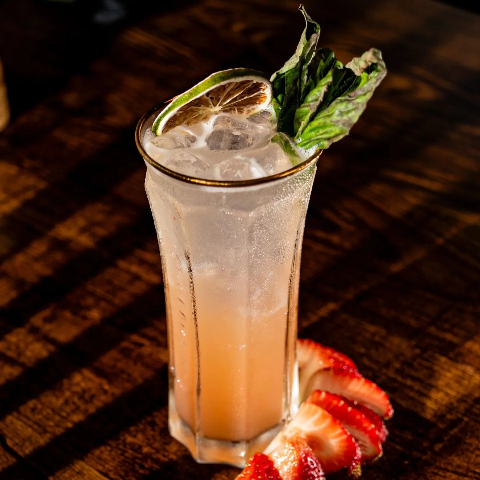 A tall glass of a pinkish cocktail garnished with a lemon or lime slice, a leaf of basil or mint, and sliced strawberries on the side, sitting on a dark wooden surface.