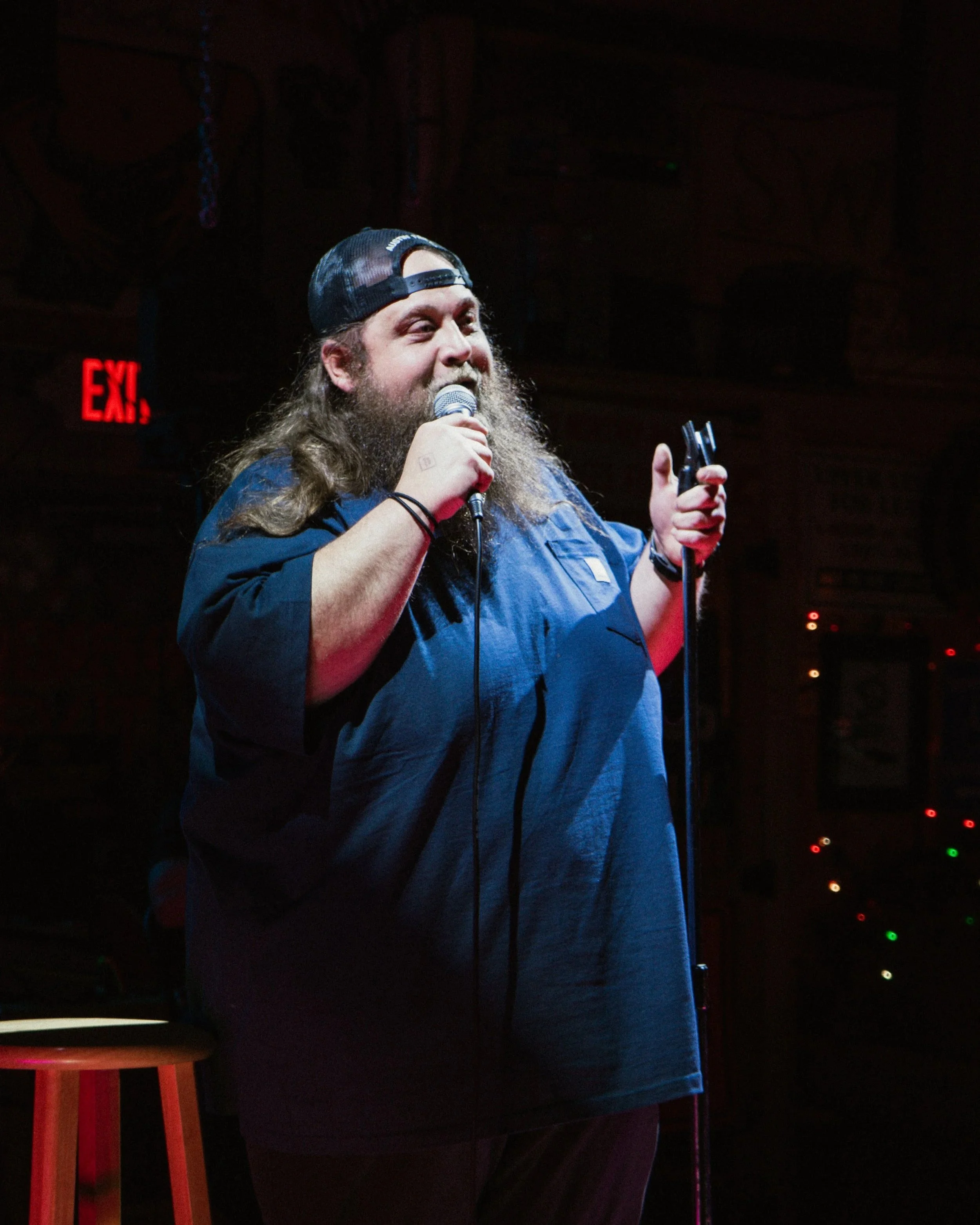 A man with a beard and long hair, wearing a backwards baseball cap and a dark shirt, speaking into a microphone on stage with a dark background and some colored lights.