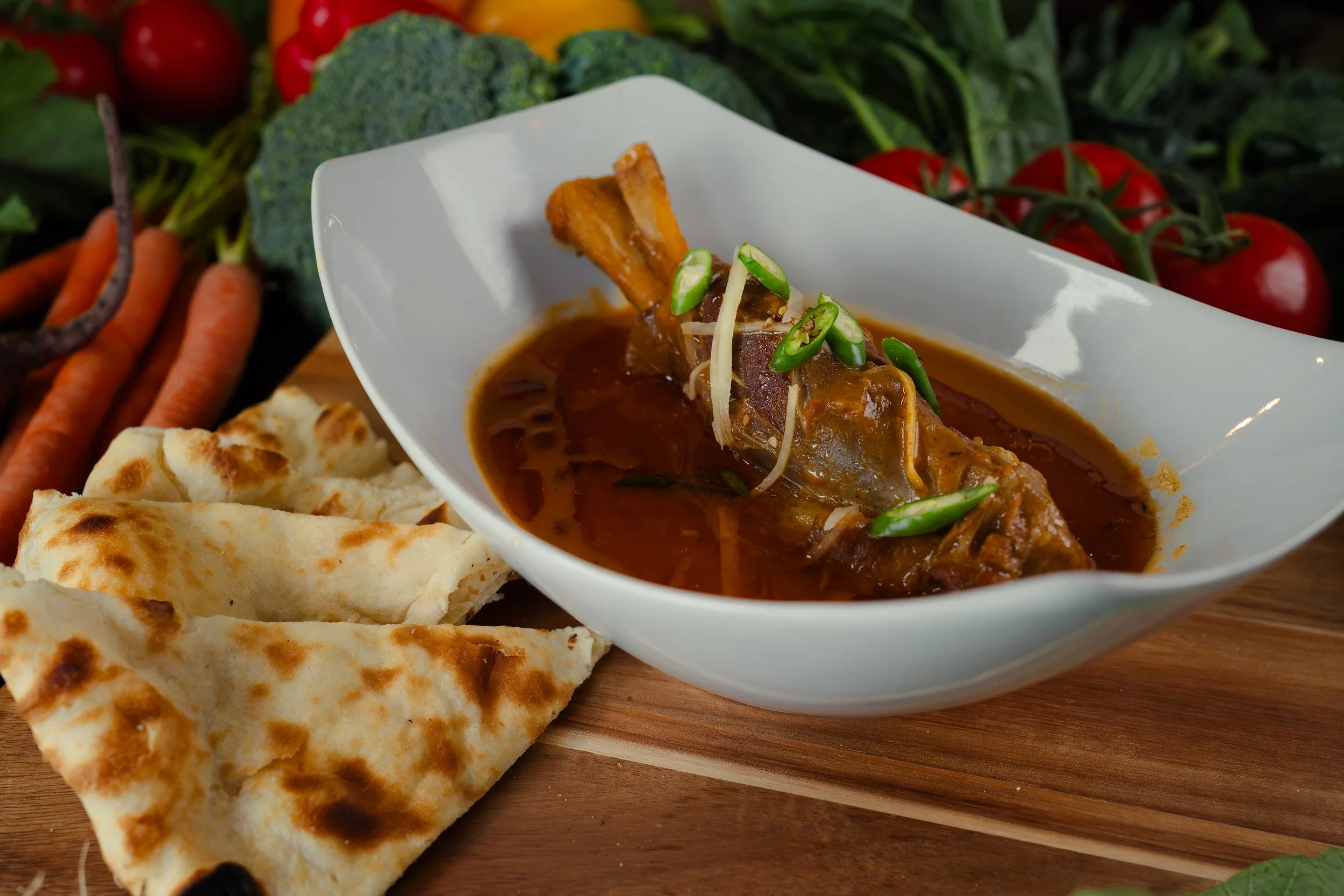 A bowl of beef stew garnished with sliced green chili peppers, accompanied by naan bread, with fresh tomatoes, broccoli, carrots, and other vegetables in the background.