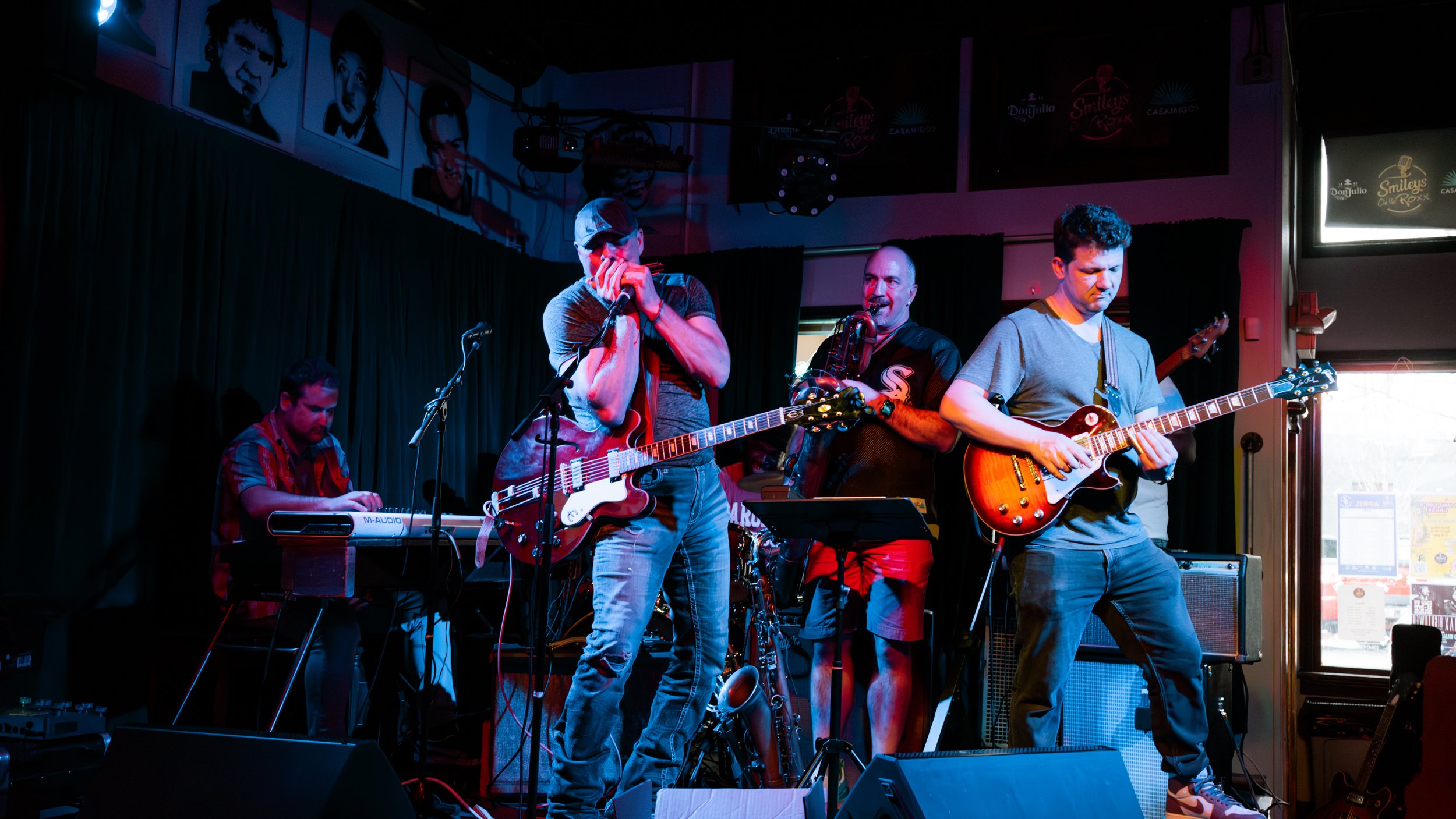 A band performing on stage with four members: a keyboard player, a vocalist with a guitar, a saxophonist, and a guitarist. They are under dim stage lighting, with a dark curtain backdrop and artwork on the walls.