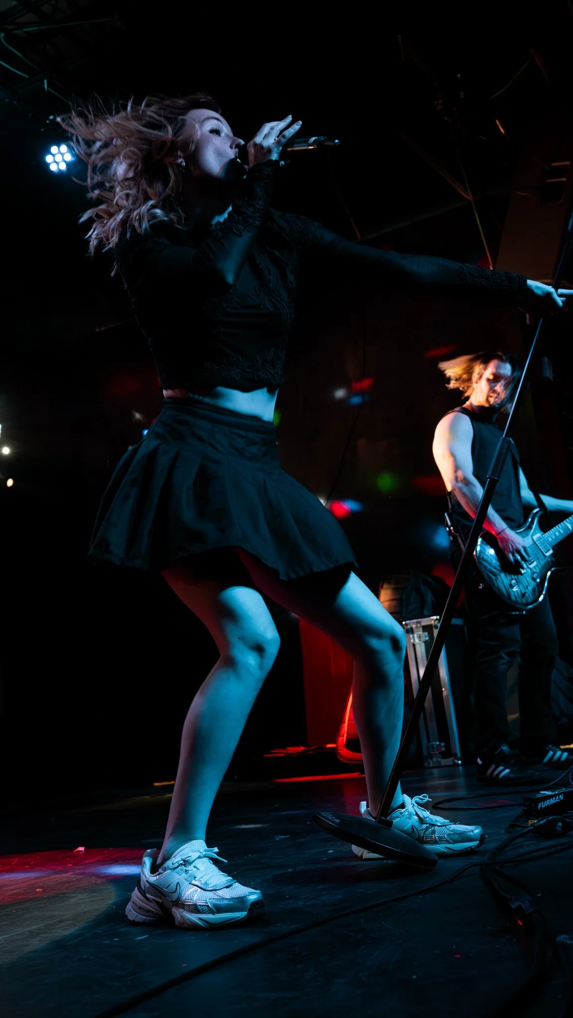 A woman singing into a microphone and a man playing an electric guitar on stage during a live performance, illuminated by stage lights.