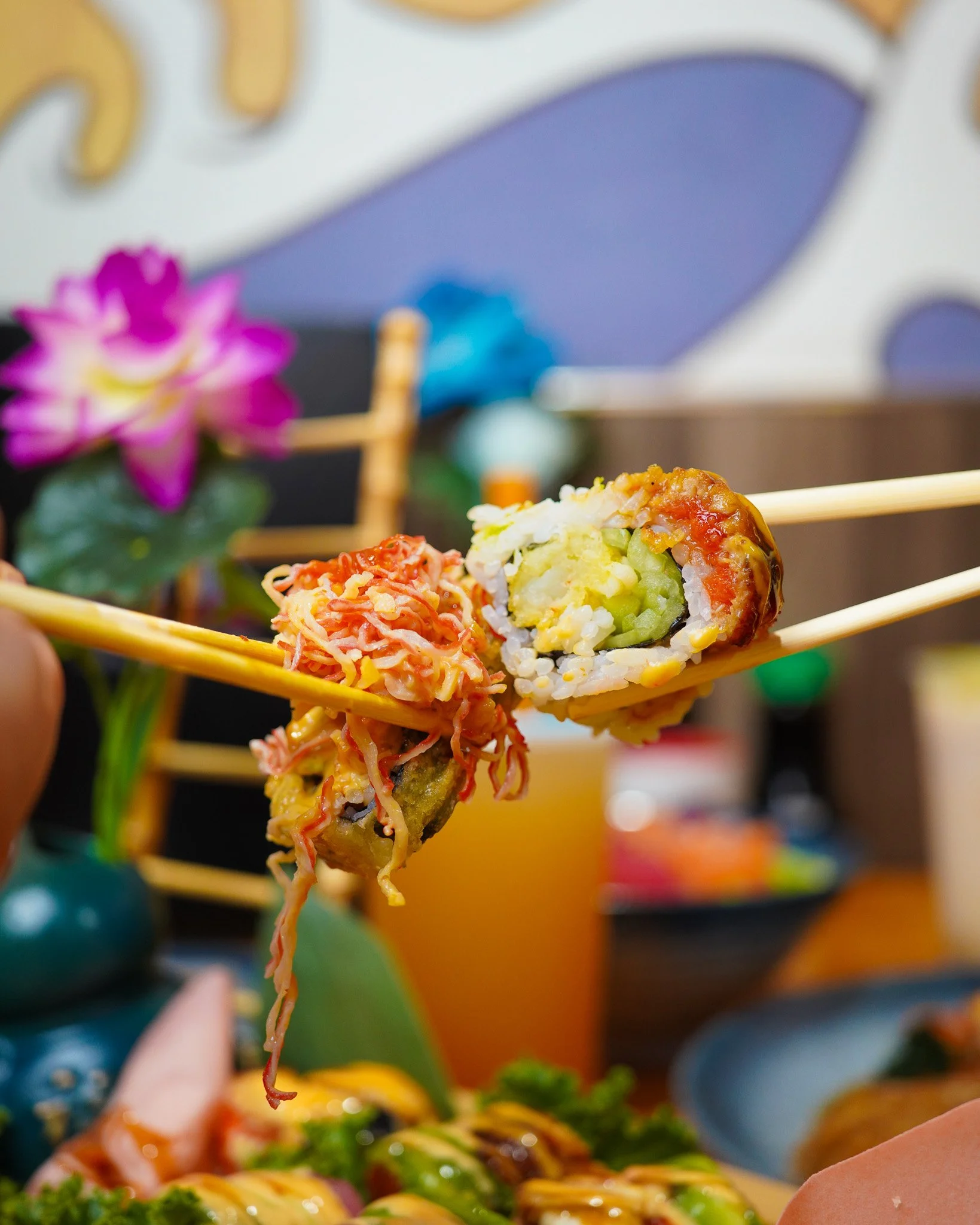 Close-up of a hand holding yellow chopsticks picking up a sushi roll with rice, vegetables, and fish, with a blurred background of colorful dishes and a purple flower.