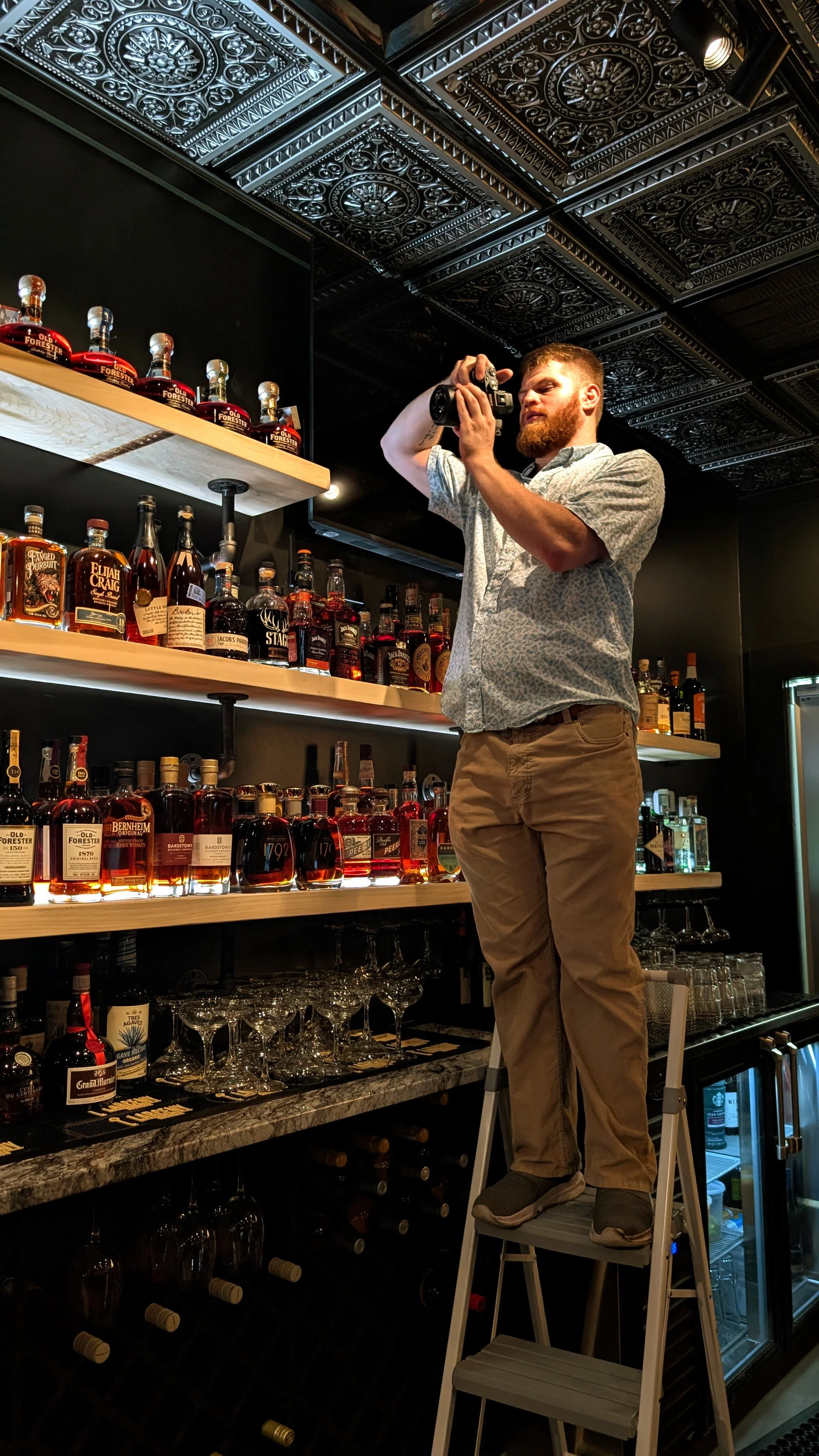 A man with a beard standing on a step ladder, taking a photo of alcohol bottles on shelves in a bar. The shelves are filled with various bottles of liquor, and the ceiling has ornate black and silver decorative panels.
