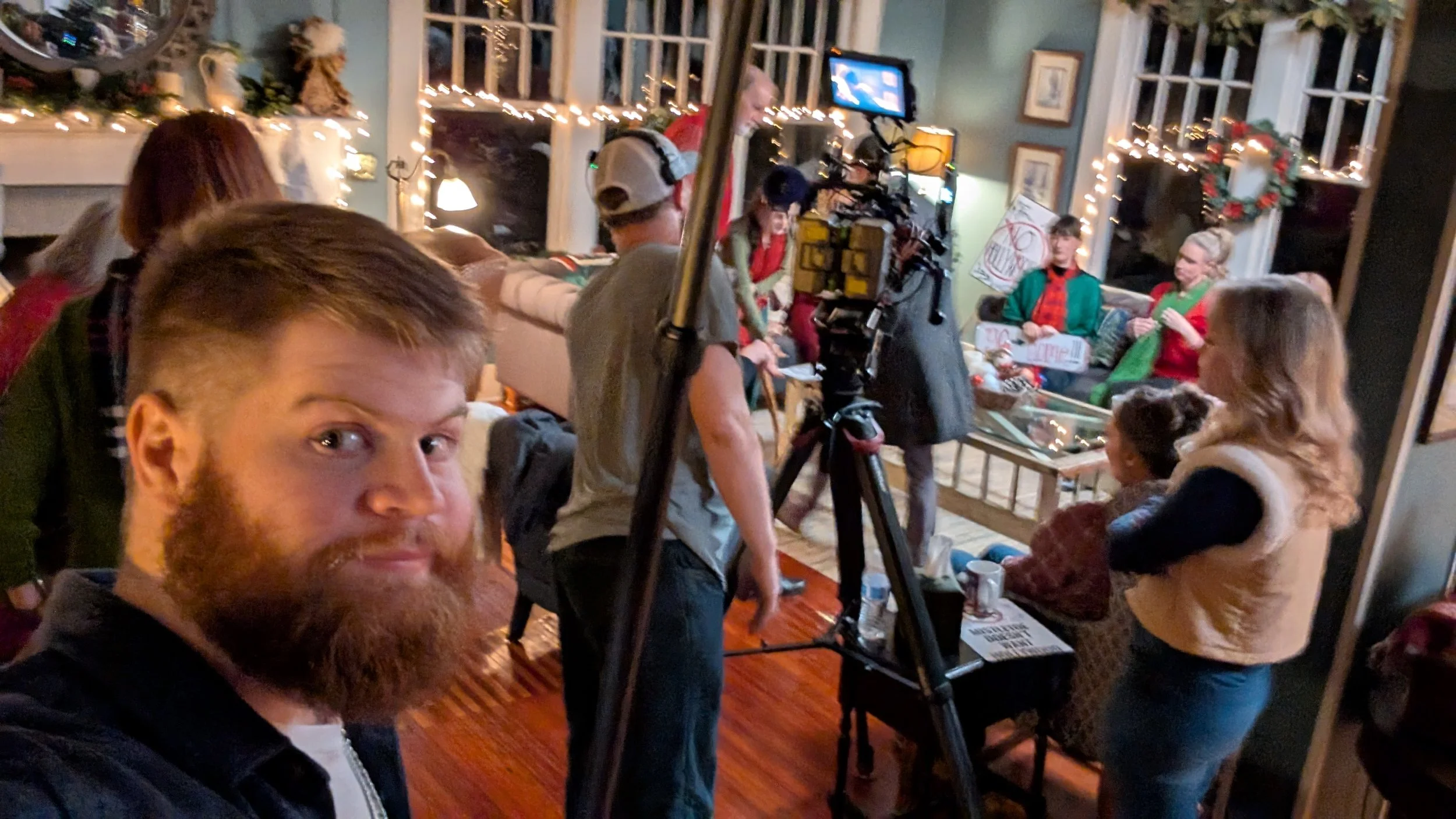 People gathered inside a festive decorated room, with string lights and holiday ornaments; some individuals are seated while others are standing or operating camera equipment.