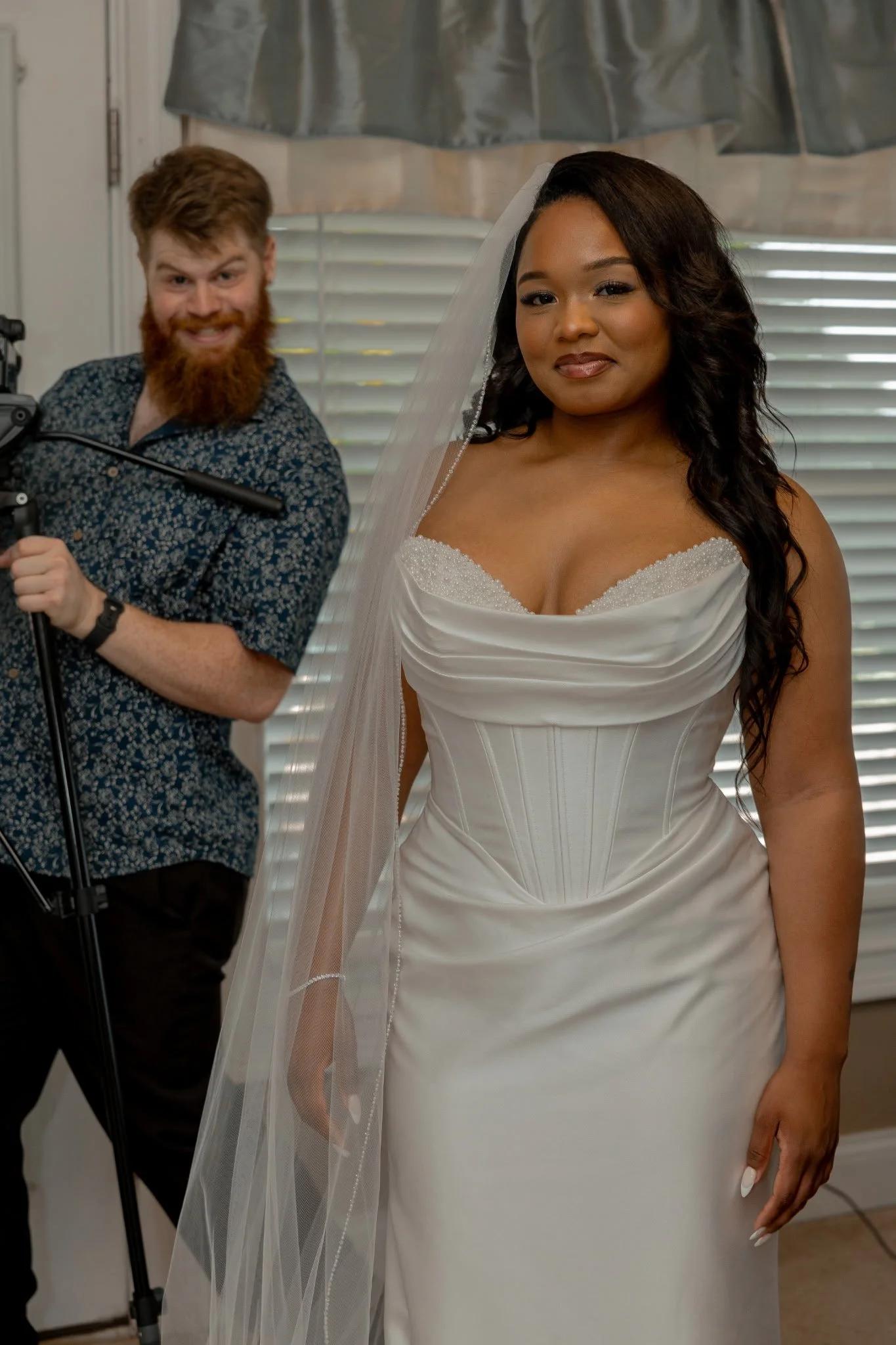 A bride in a white wedding dress with a veil standing inside a room with blinds, beside a man with red hair and a beard, wearing a patterned shirt, smiling.