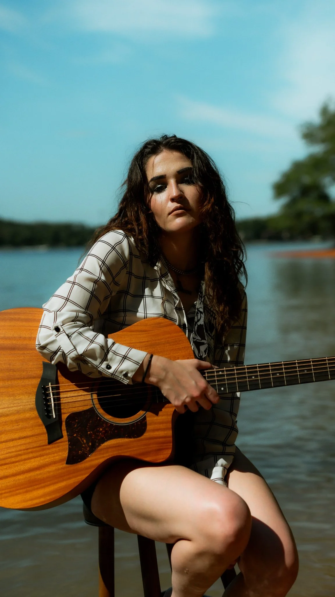 A woman with long dark hair sitting outdoors near a body of water, holding an acoustic guitar, wearing a checkered shirt, with trees and a blue sky in the background.