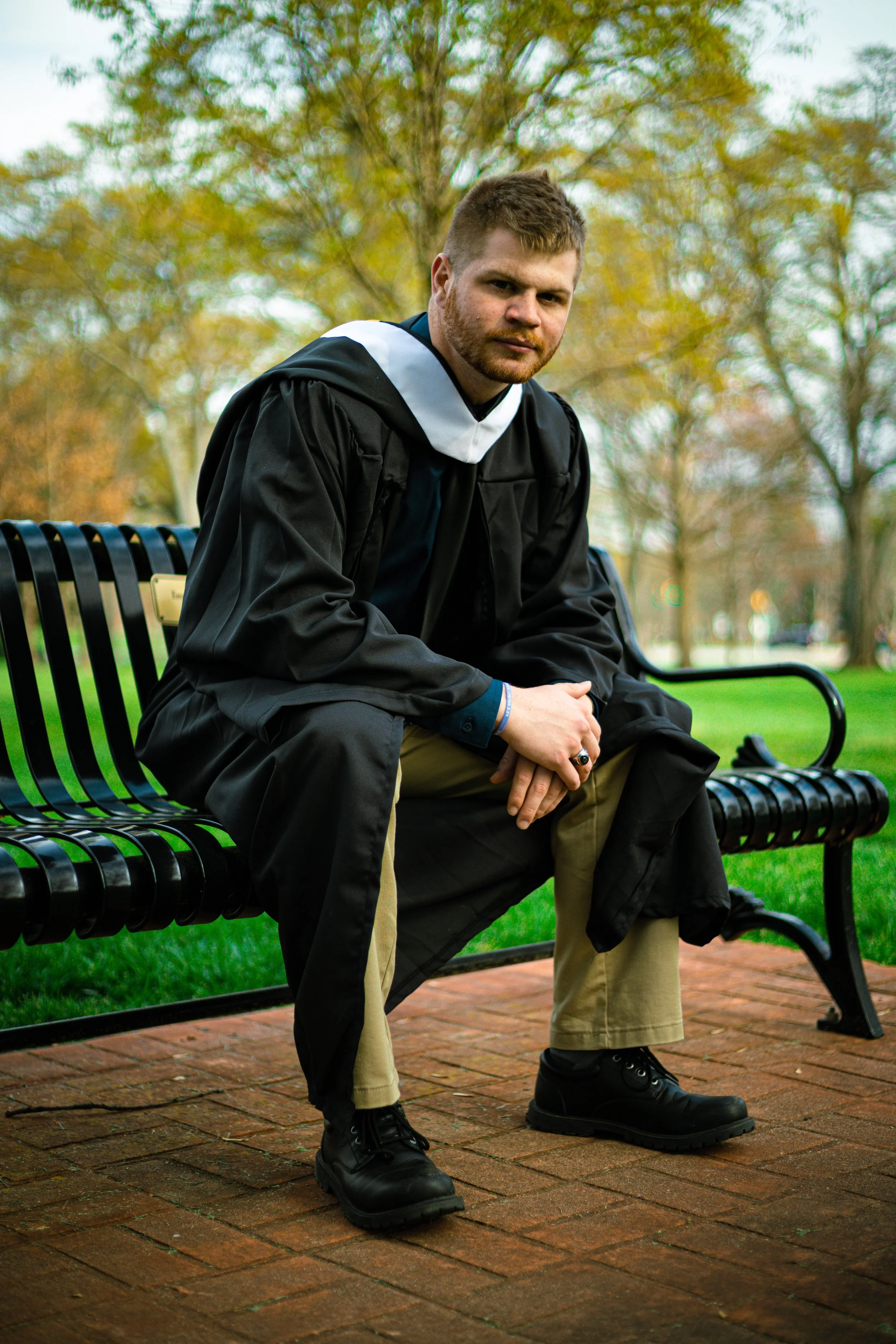 A young man with a beard and short hair sitting on a park bench, dressed in a black graduation gown and cap. He is outdoors with trees showing fall foliage in the background, sitting with his hands clasped, wearing beige pants and black boots.