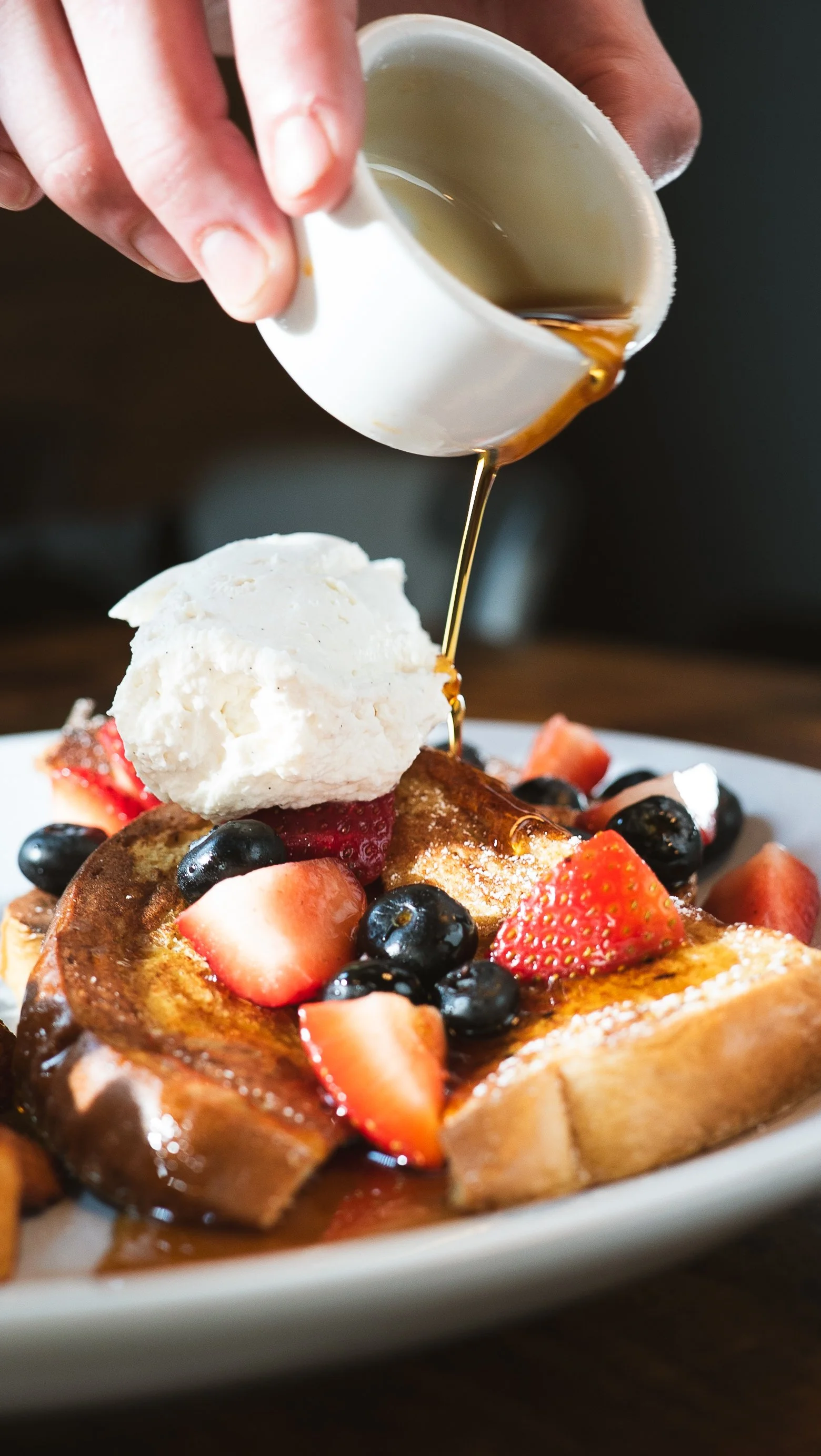 A plate of French toast topped with sliced strawberries and blueberries, with a large scoop of vanilla ice cream, and syrup being poured over it.