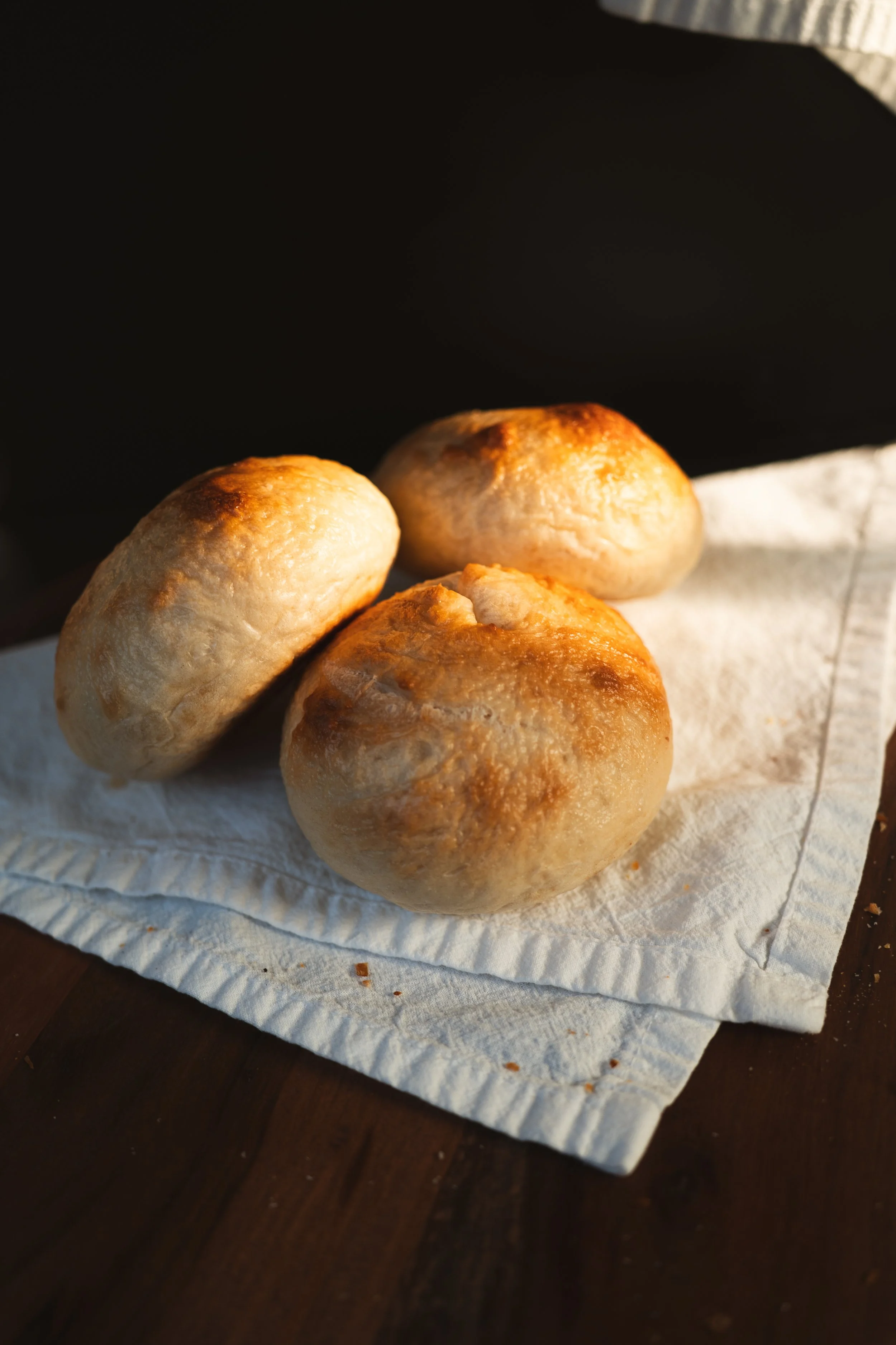 Three freshly baked bread rolls on a piece of parchment paper, placed on a dark wooden surface.
