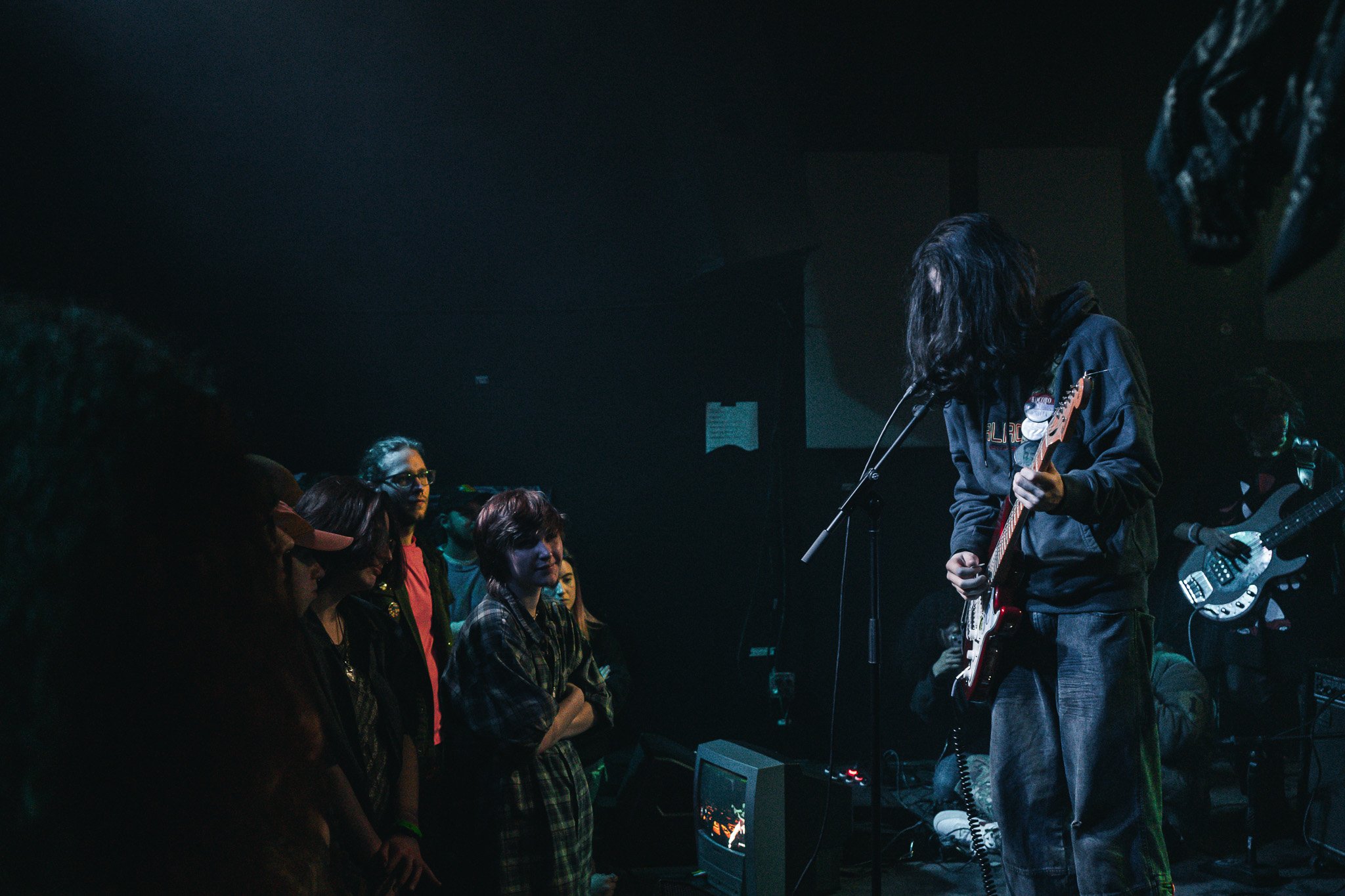 A musician with long hair playing an electric guitar on stage during a live performance while an audience watches in the dark. The stage is dimly lit.