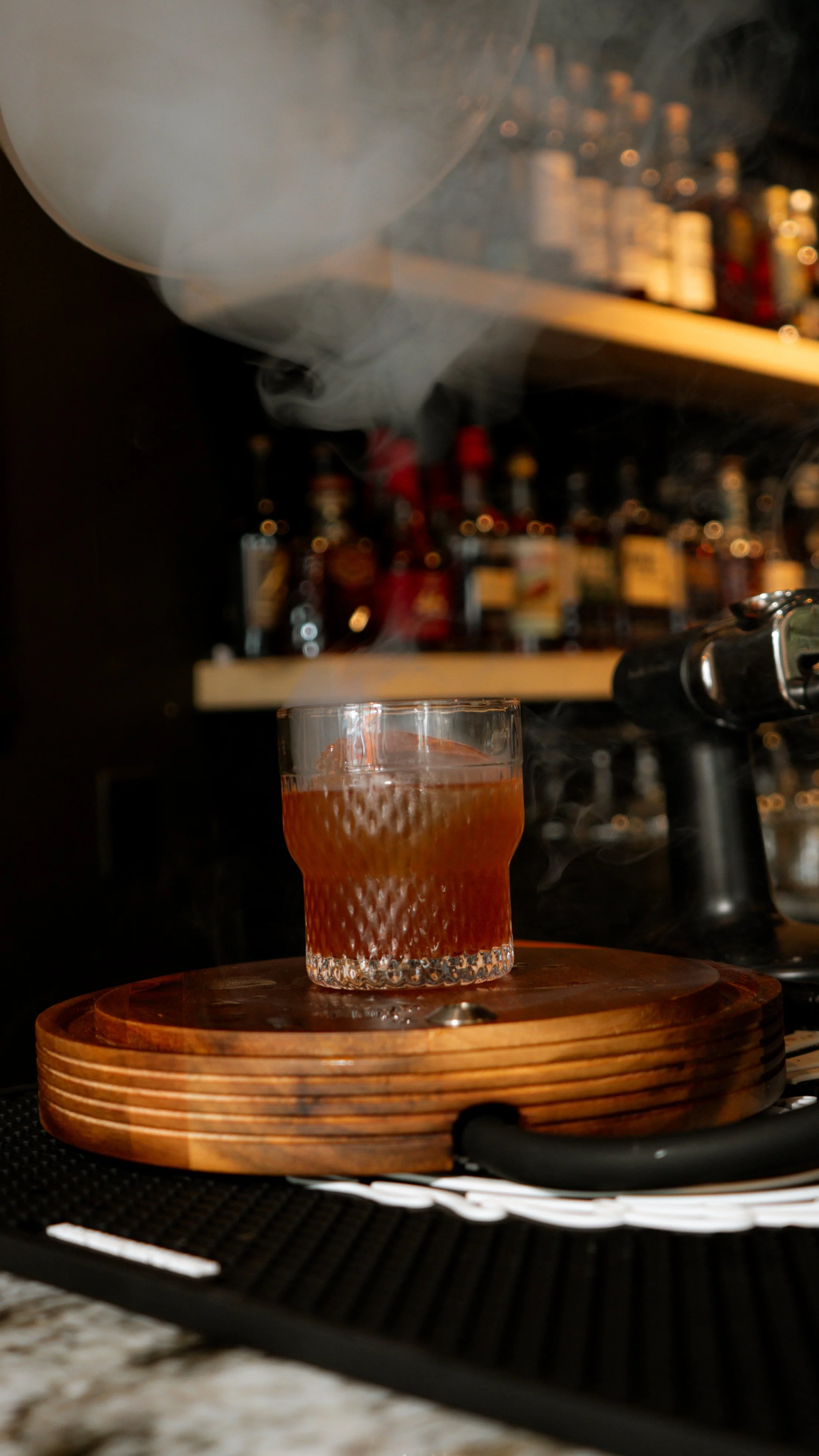 A glass with a textured pattern on the outside, filled with a cocktail, sits on a round wooden tray on a bar counter, with smoke or vapor rising from the drink.