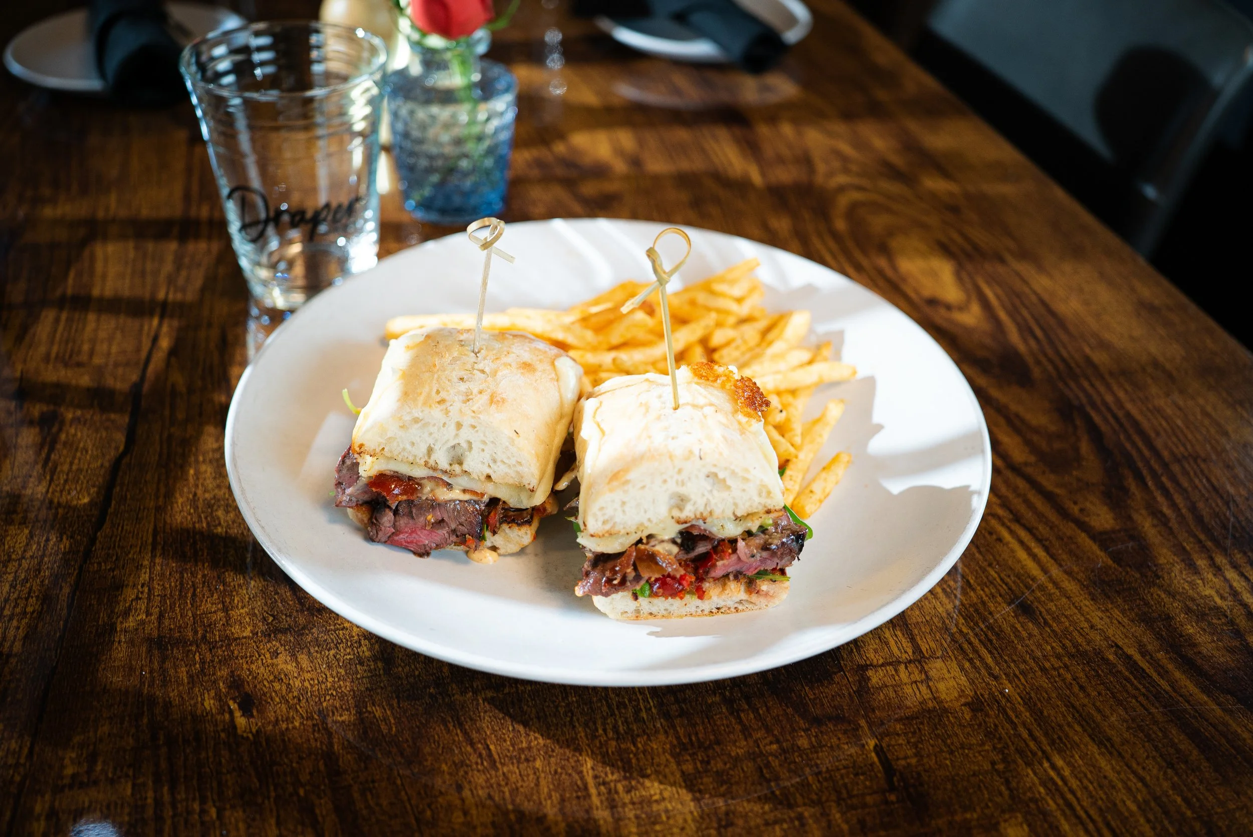 Plate with a half-cut sandwich and French fries, two glasses of water, and a small flower in a vase on a wooden table.