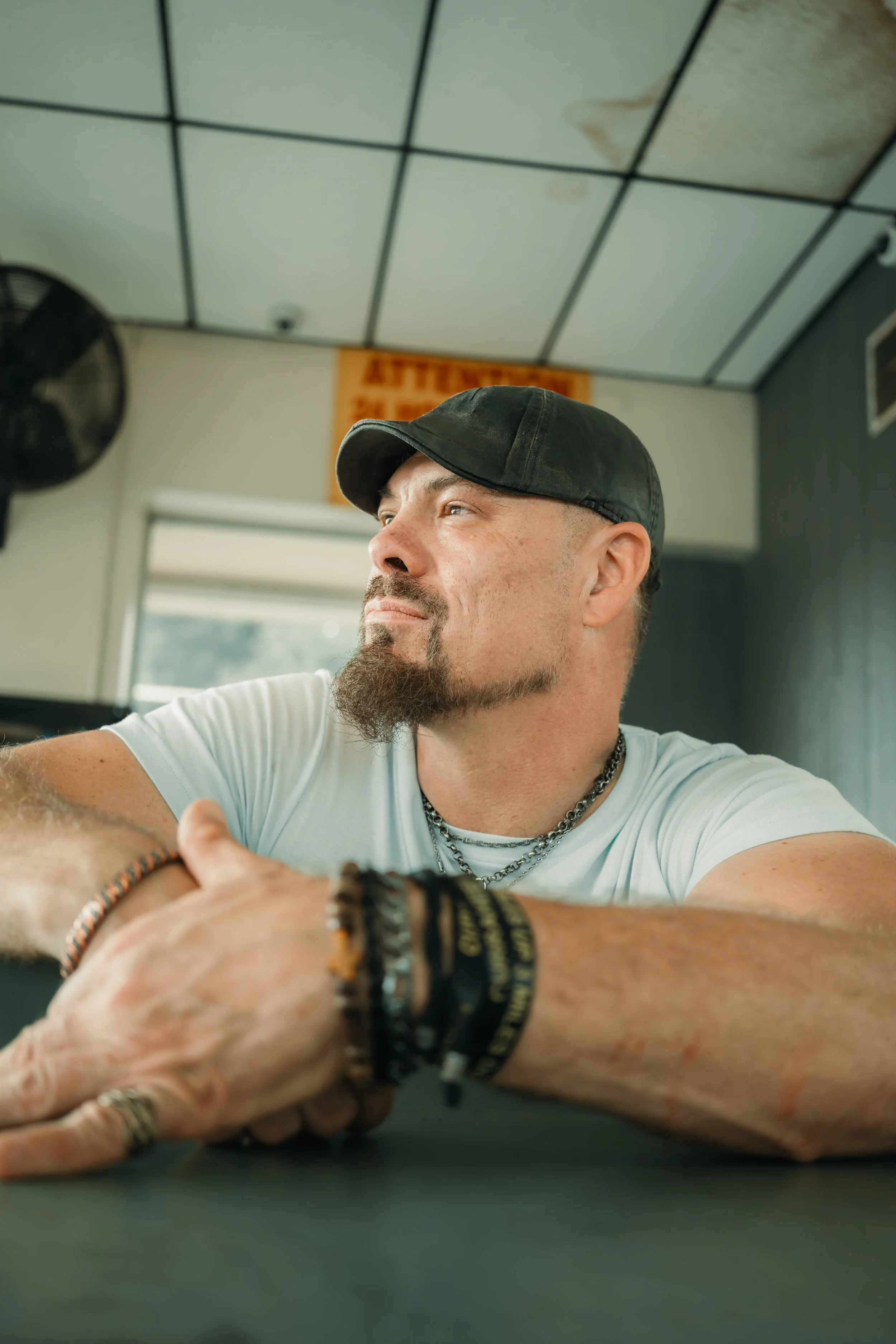 A man with a beard, wearing a black cap, white t-shirt, and layered necklaces, sits at a table with his hands resting on the surface. The room has a ceiling with black grid lines, a black wall fan, and a sign in the background reading 'ATTENTION.'