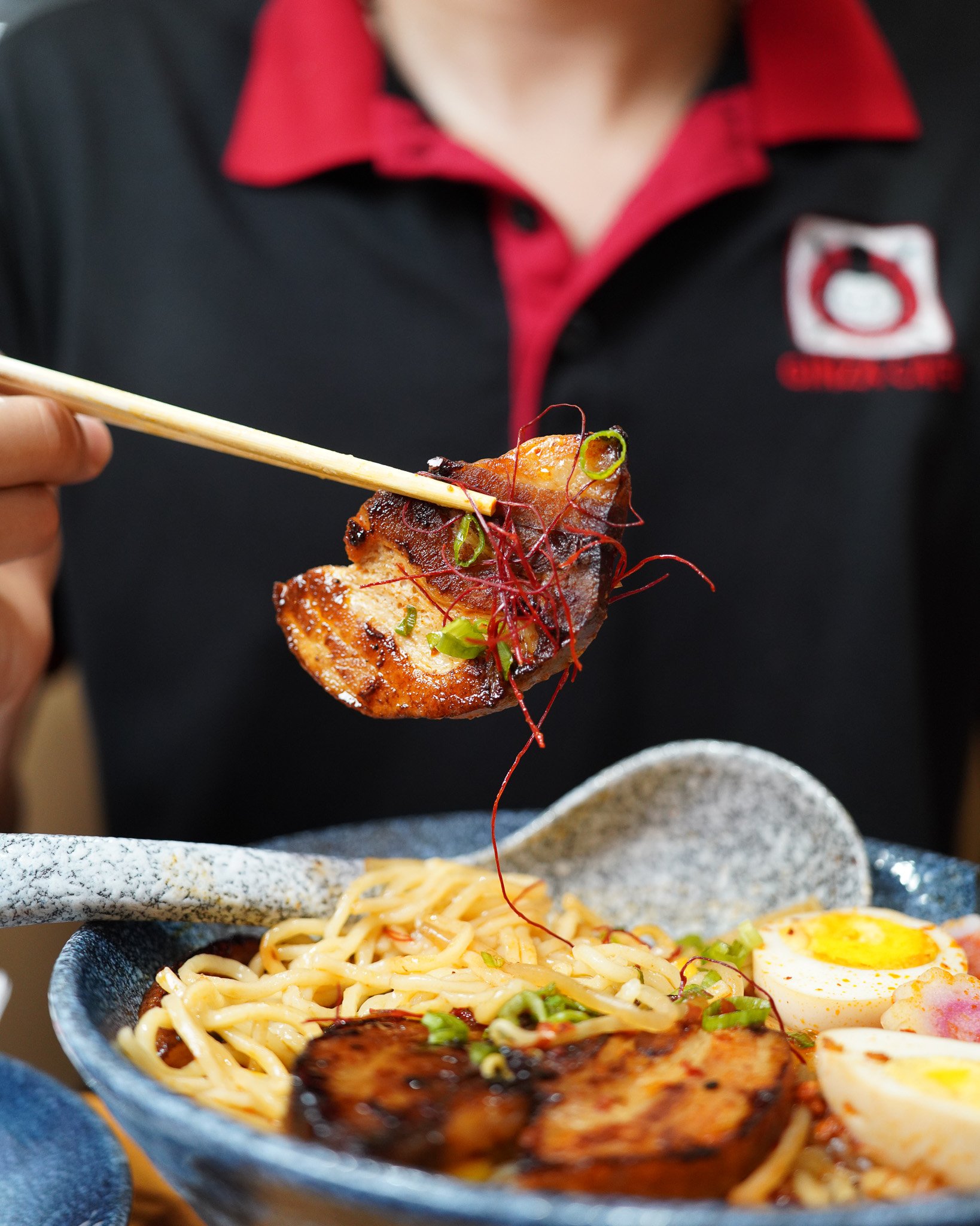 Person holding chopsticks with grilled meat over a bowl of ramen noodles topped with green onions, soft boiled egg, and slices of meat, with a second bowl of noodles visible in the background.