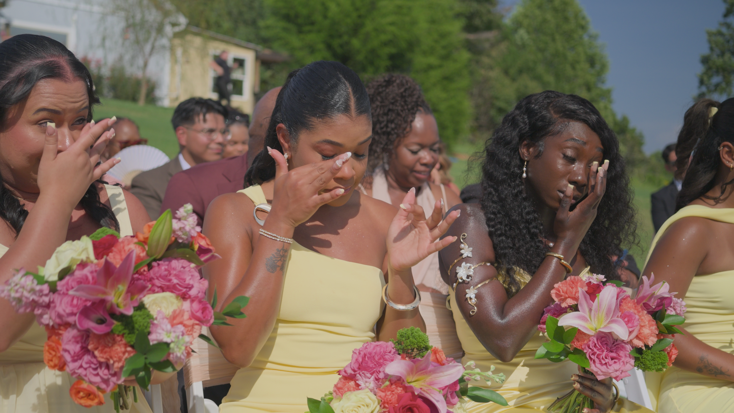 Group of women crying at an outdoor wedding ceremony, holding flowers, with greenery and blue sky in the background.