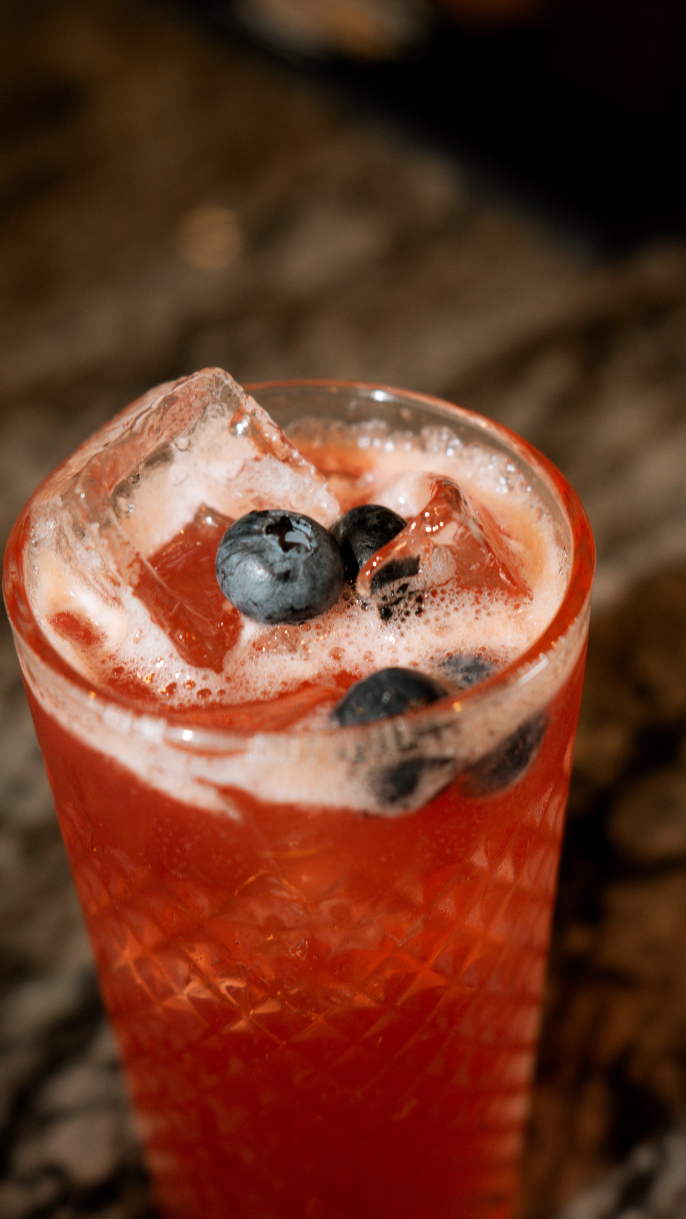 Close-up of a tall glass filled with a pink or reddish beverage, ice cubes, and blueberries on top, placed on a dark, textured surface.