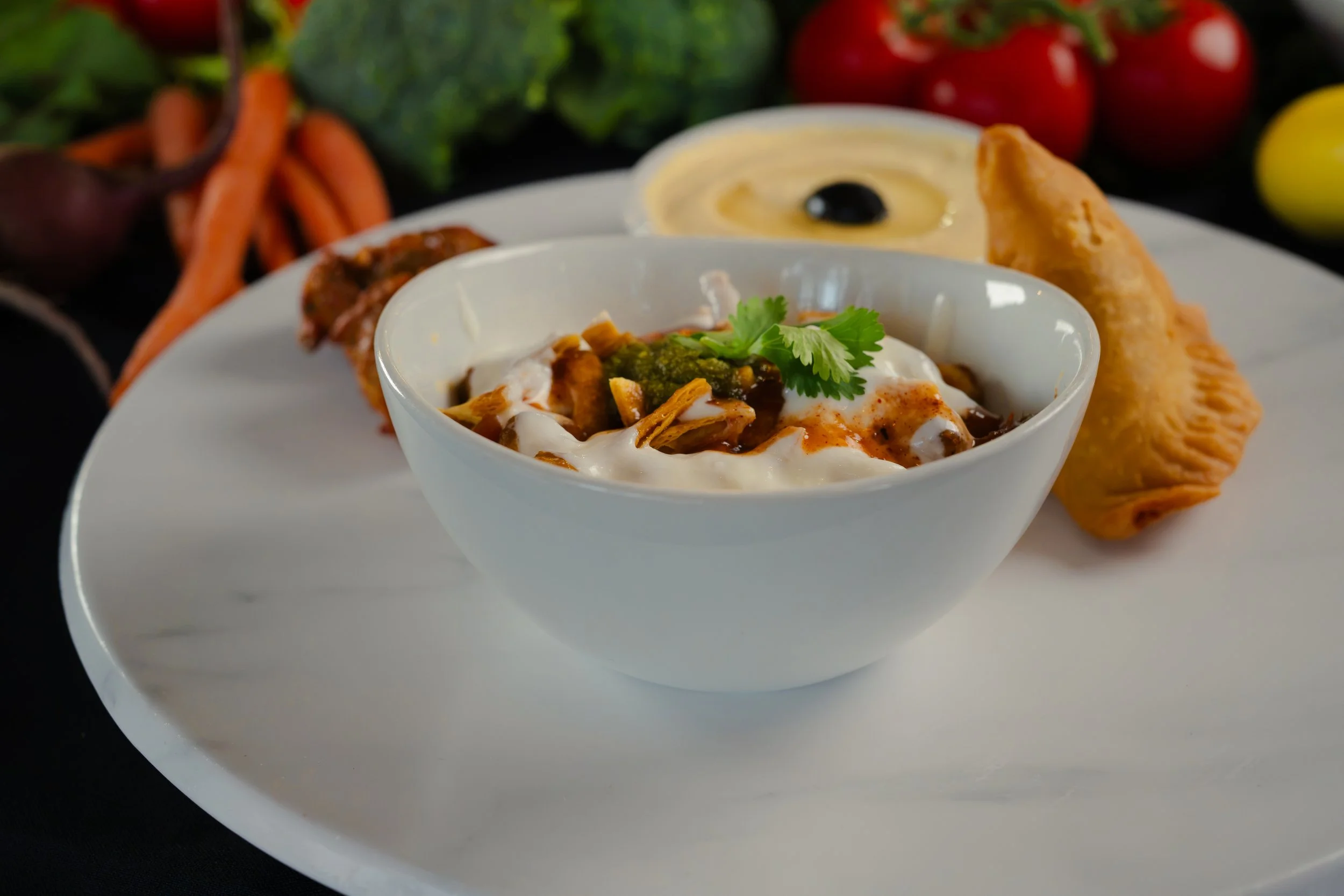 A white bowl of Indian chicken curry garnished with cilantro and served with naan bread, accompanied by a side of raita, on a white plate with vegetables in the background.