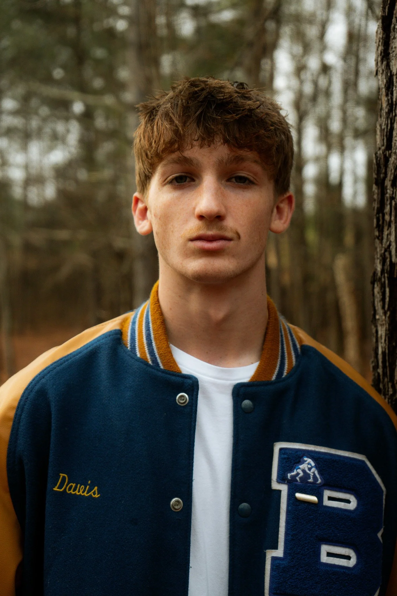 A young man with curly brown hair and light skin standing outdoors in a wooded area.
