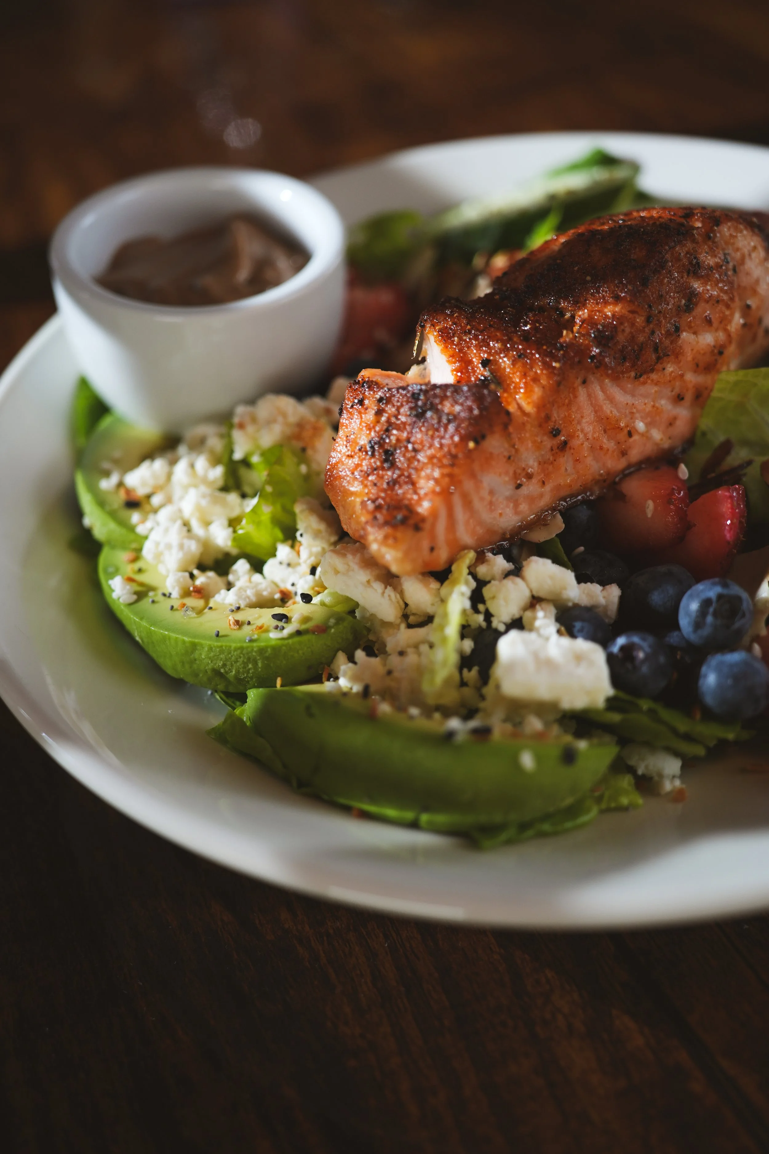 A white plate with grilled salmon, avocado slices, crumbled cheese, mixed berries, and a small bowl of gravy or sauce, set on a dark wooden surface.