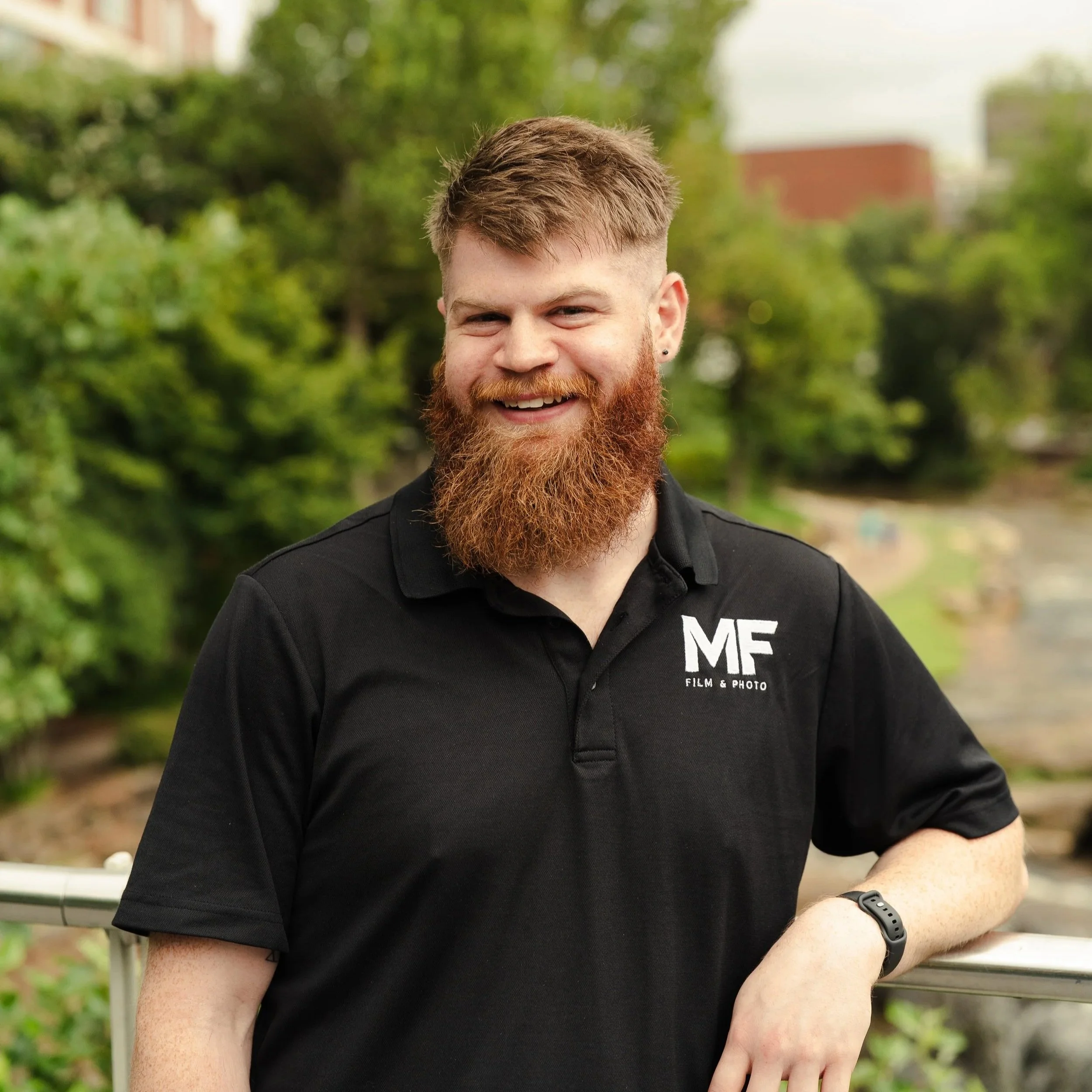 Close-up of a smiling man with red hair, beard, and earrings wearing a black shirt with white text, standing outdoors with green trees in the background.