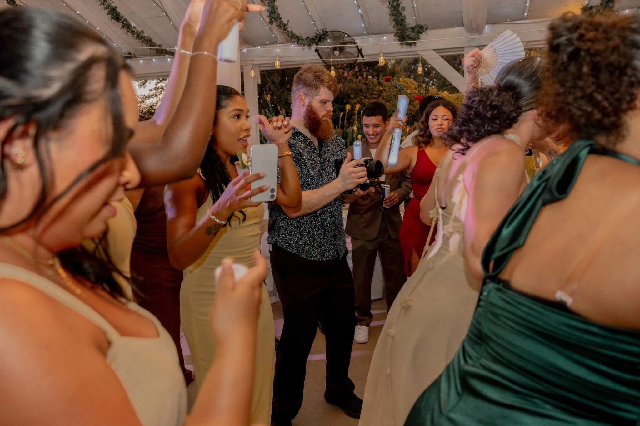 Group of diverse people celebrating at a party, raising glasses, taking photos, and smiling under string lights and greenery with a man with red hair and a beard filming in the middle.