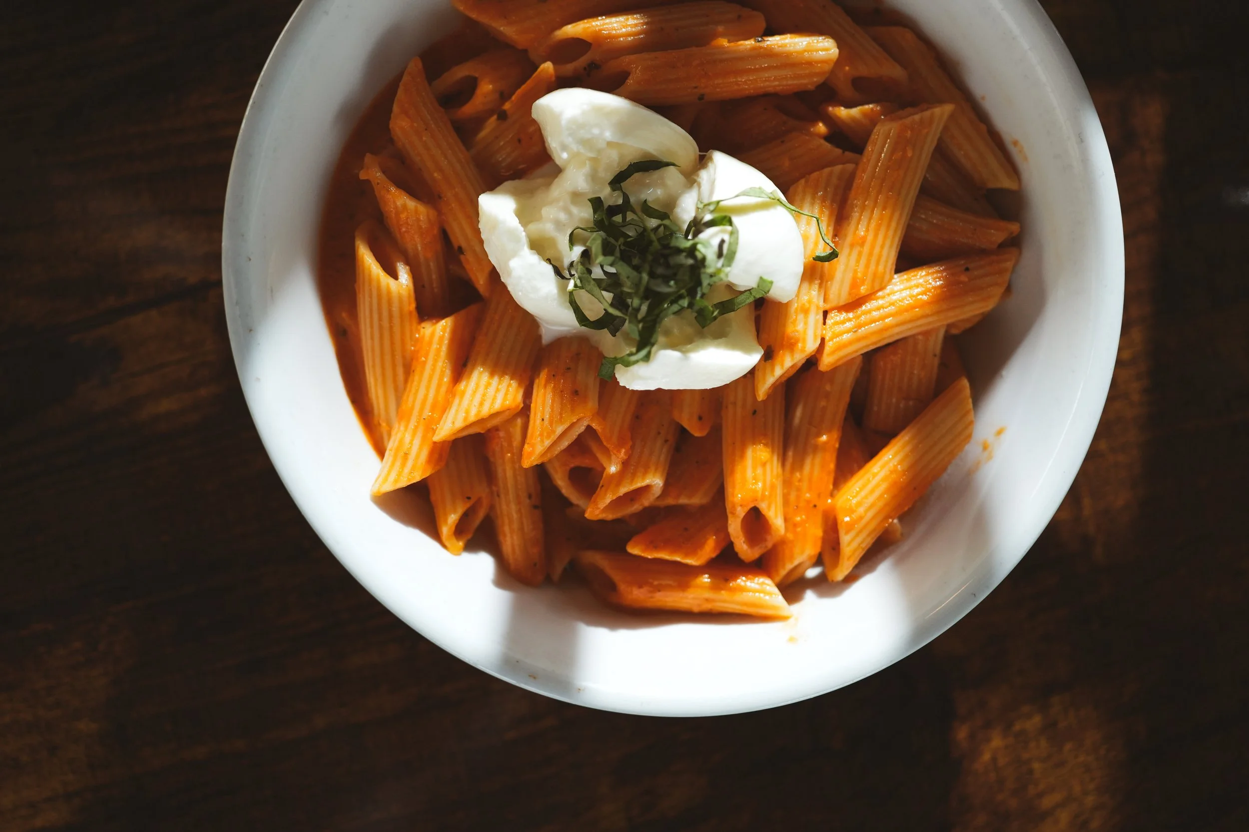 A white bowl of cooked penne pasta with orange-colored marinara sauce, topped with a dollop of sour cream or ricotta cheese, garnished with chopped herbs, on a dark wooden surface.