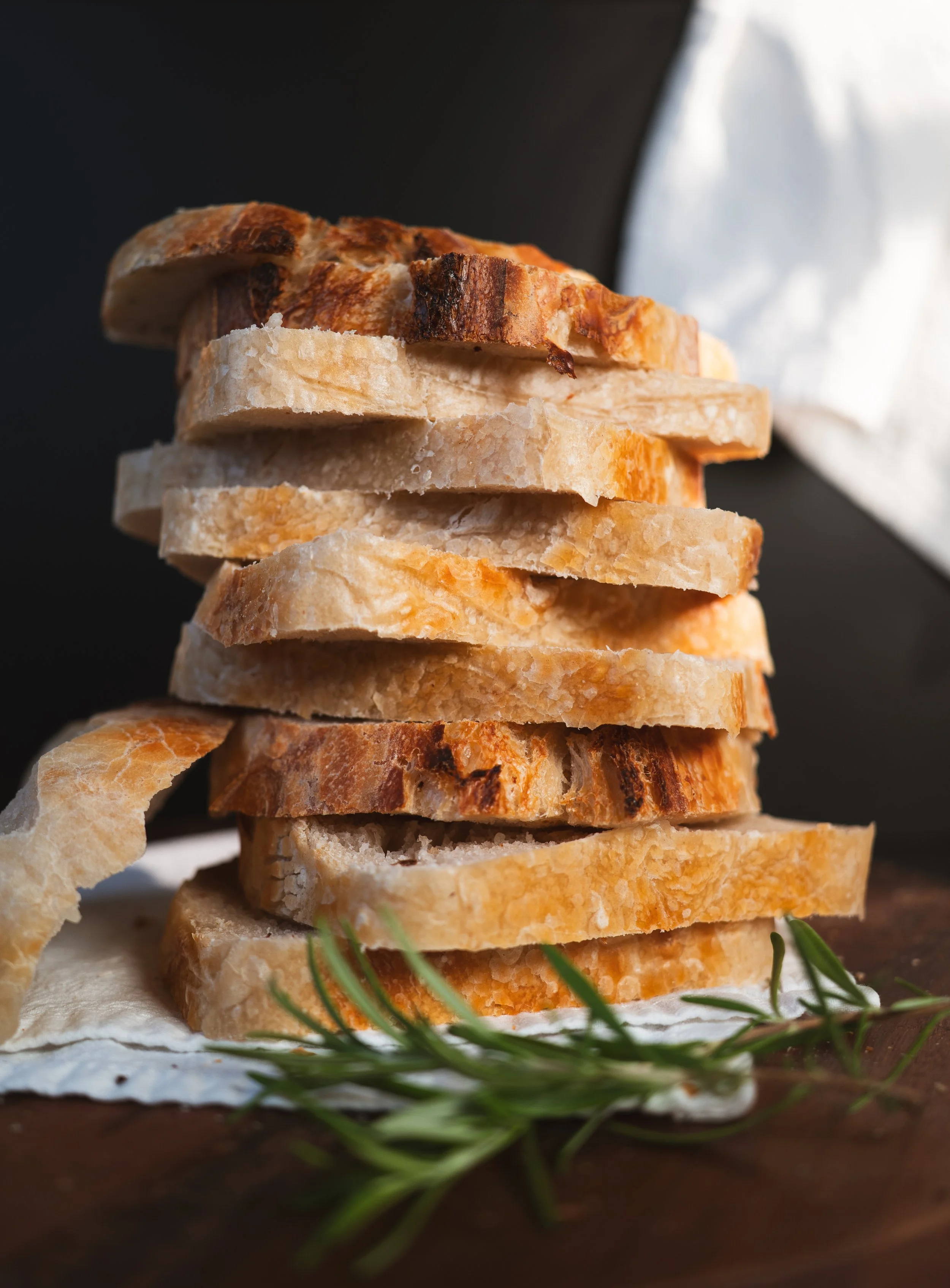 Stacked slices of toasted bread on a piece of paper, with green herbs in the foreground and a dark background.