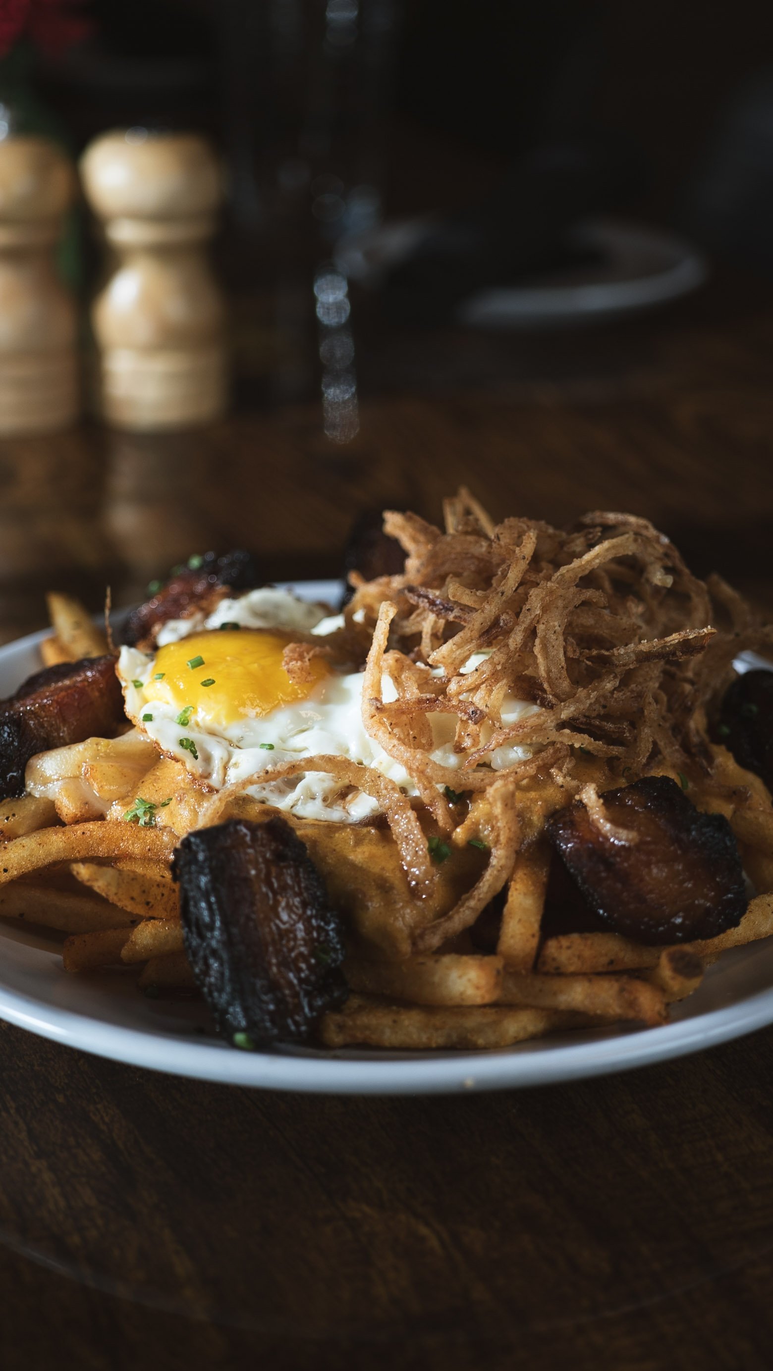 Plate of loaded fries topped with a fried egg, shredded fried onions, and chunks of pulled pork, on a dark wooden table with salt and pepper shakers in the background.