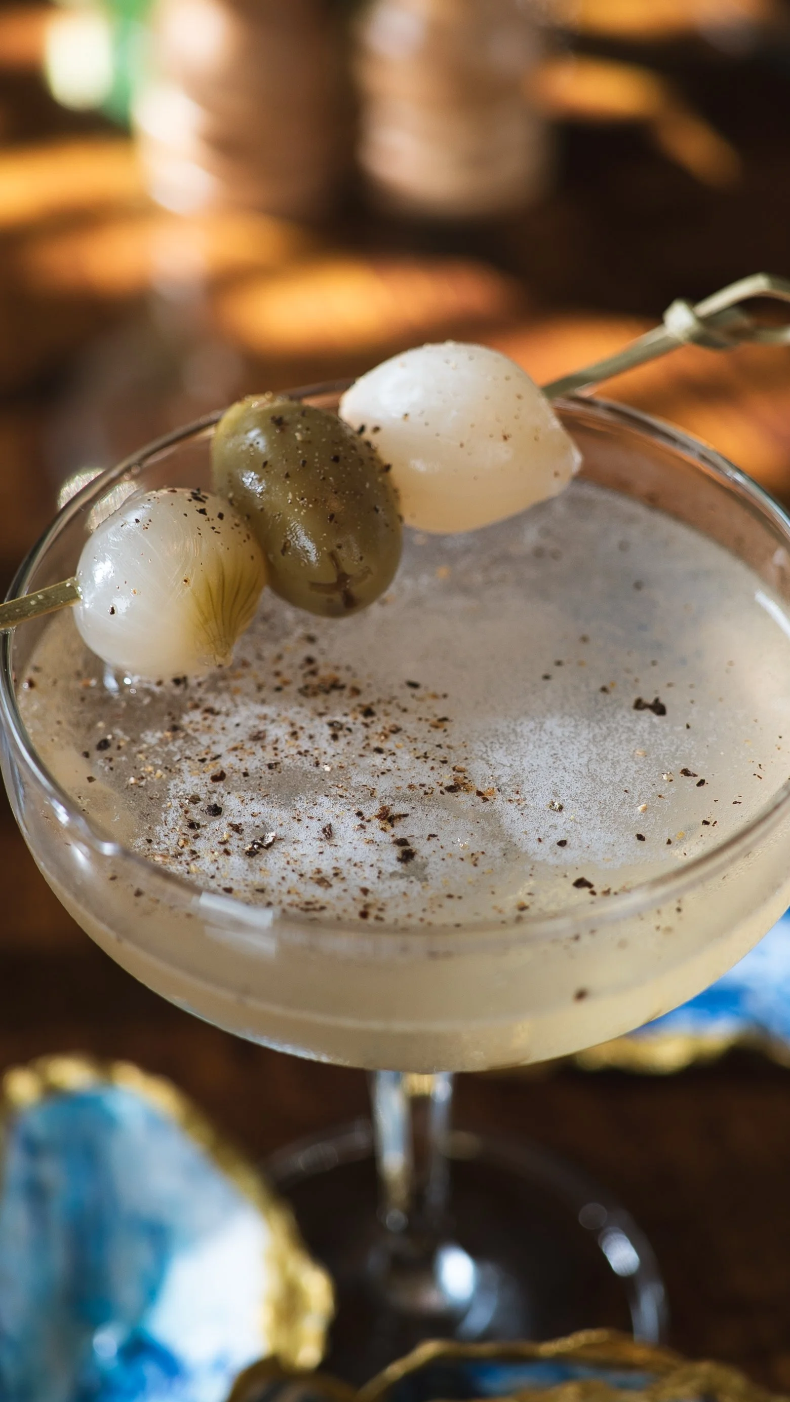 A cocktail drink garnished with three pearl onions speared on a skewer, with a sprinkle of black pepper and spices on top, in a cocktail glass.