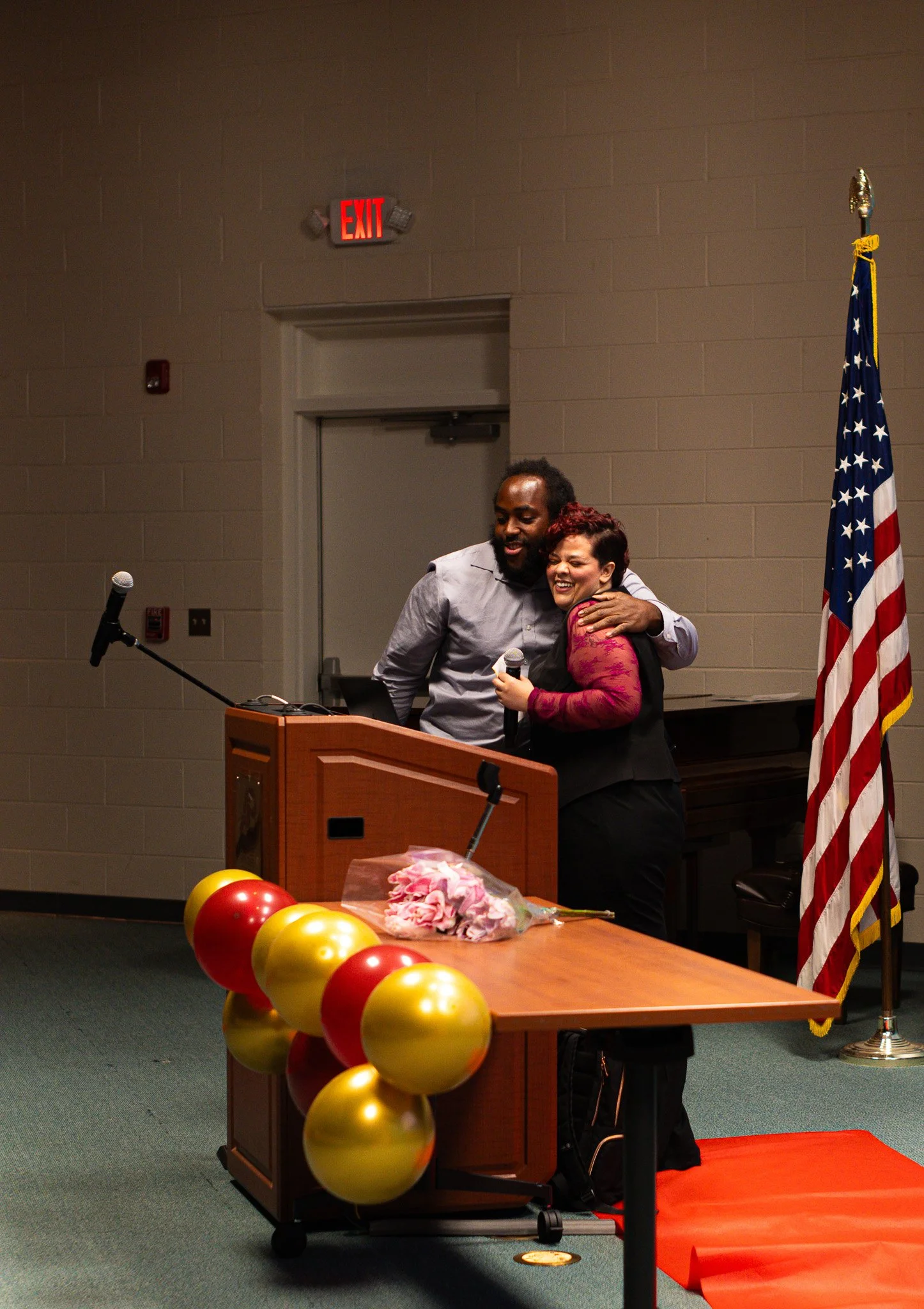 Two people hugging at the inaugural Asheville Mental Health Film Festival, with balloons and flowers on a table, American flag to the right, microphone at a podium, and exit sign in the background.