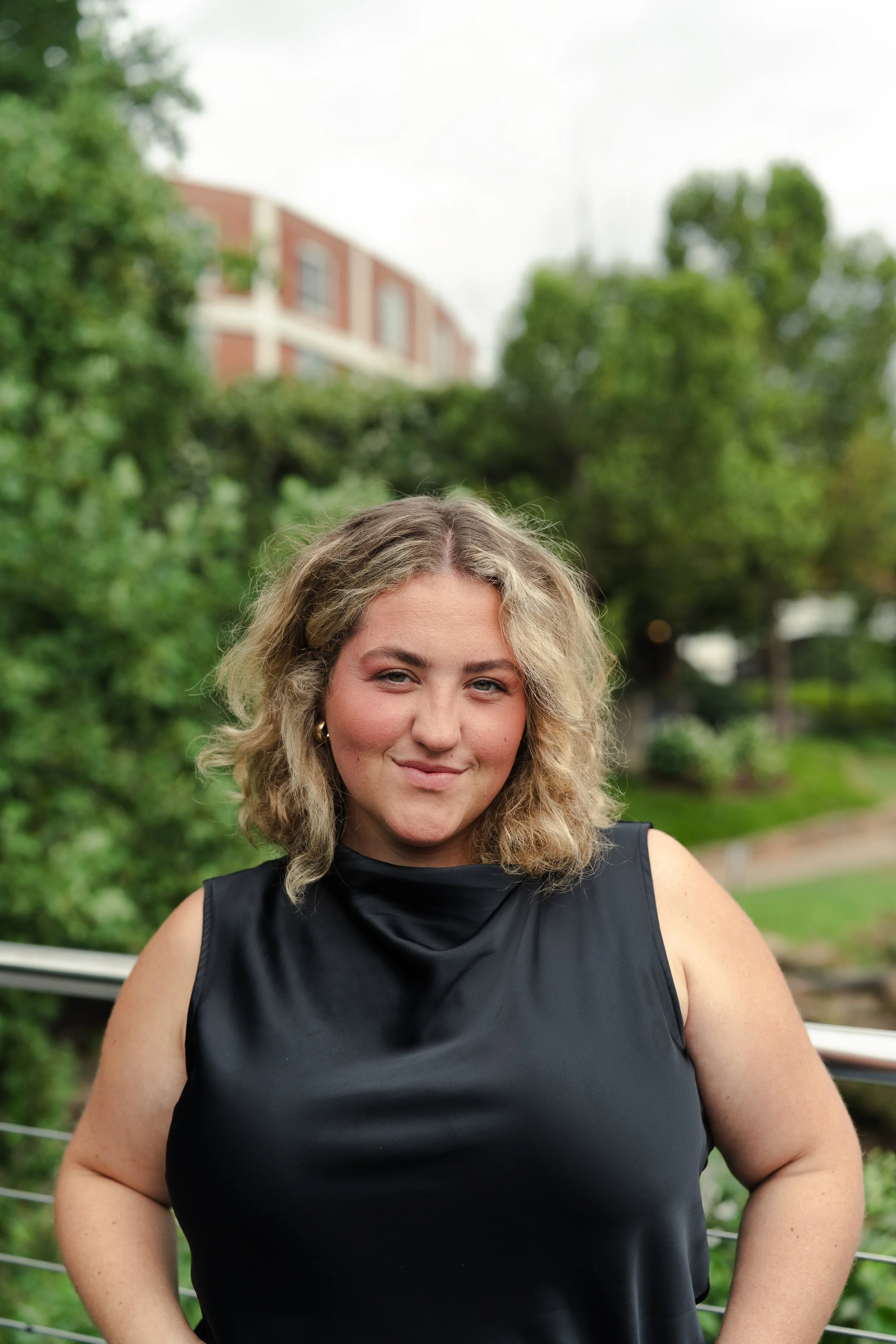 A woman with wavy blonde hair wearing a black sleeveless top standing outdoors with greenery and trees in the background.