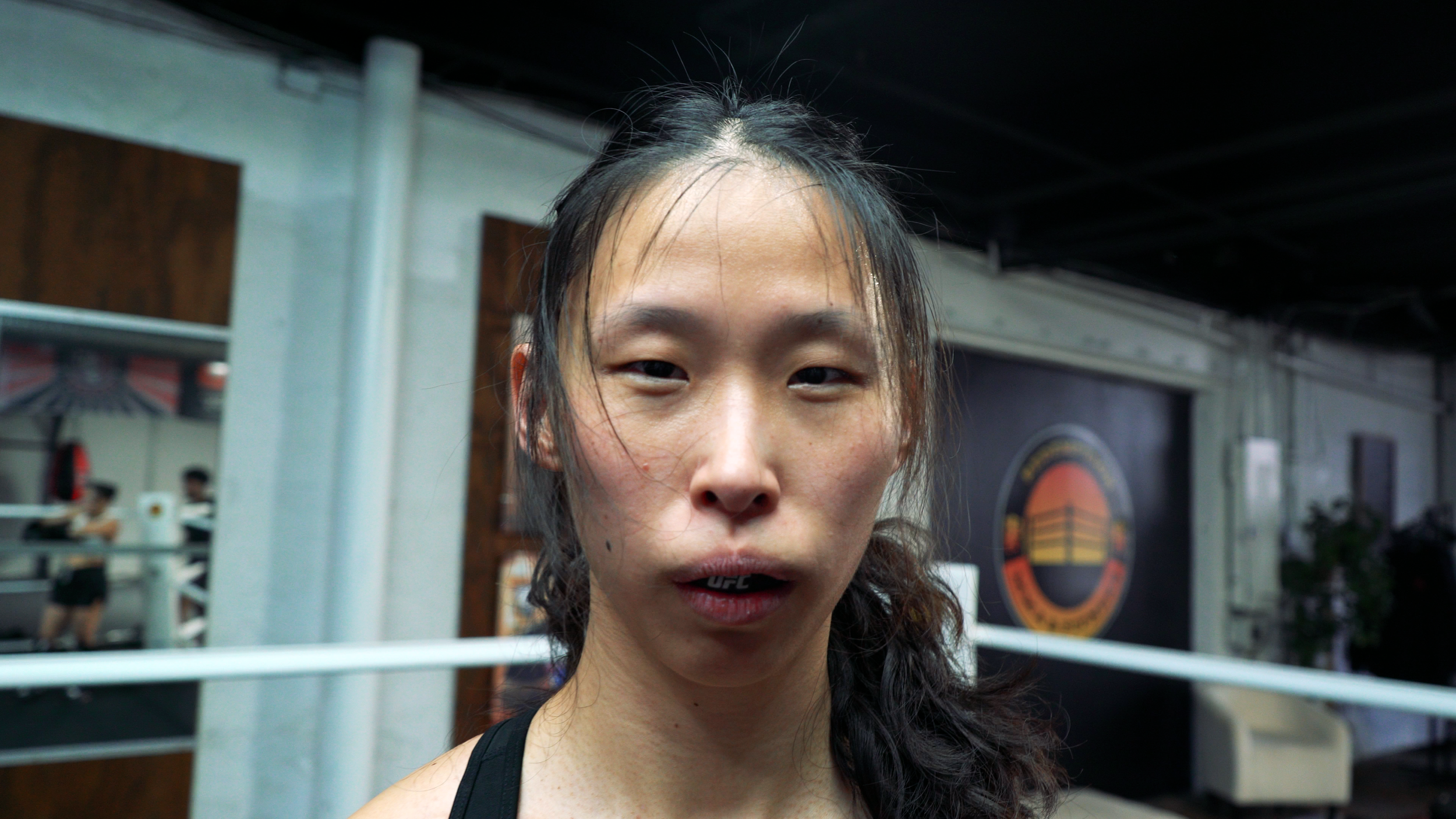 Close-up of a woman with sweaty hair in a gym or training area with boxing ring in the background.
