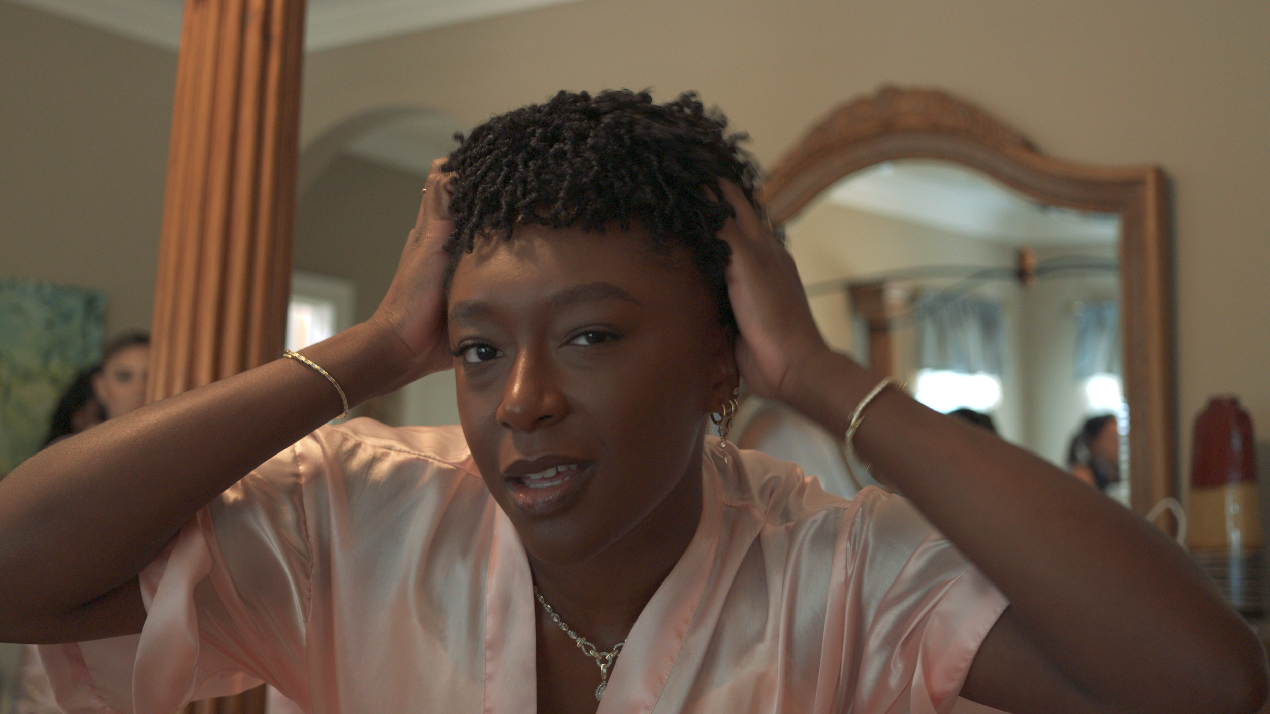 Young woman with short curly hair adjusting her hair in front of a mirror, wearing a light pink satin robe, in a warmly lit room.