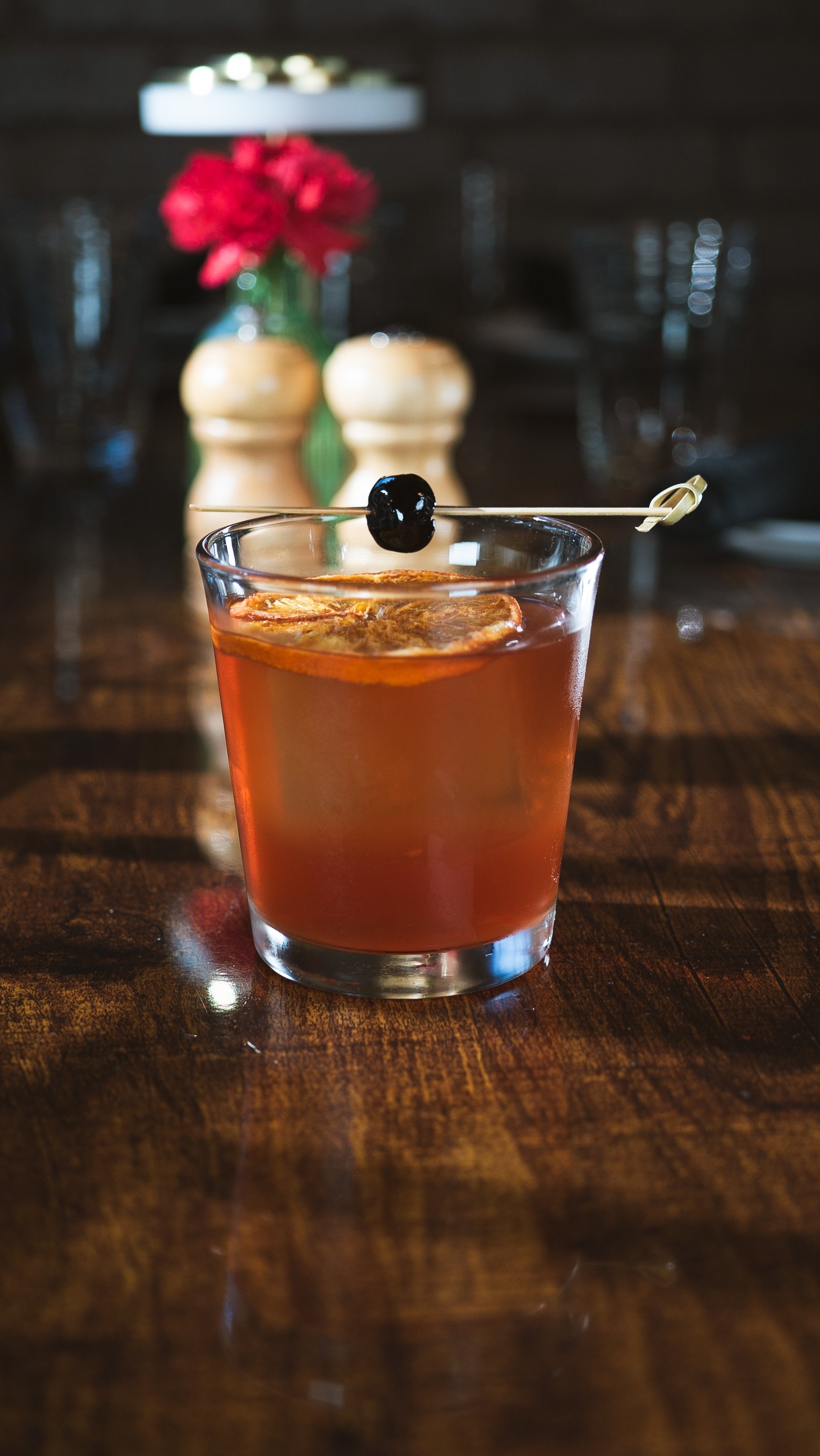 A glass of cocktail with a cherry on a skewer and a slice of citrus fruit, on a wooden table with a blurred background of a vase with red flowers.