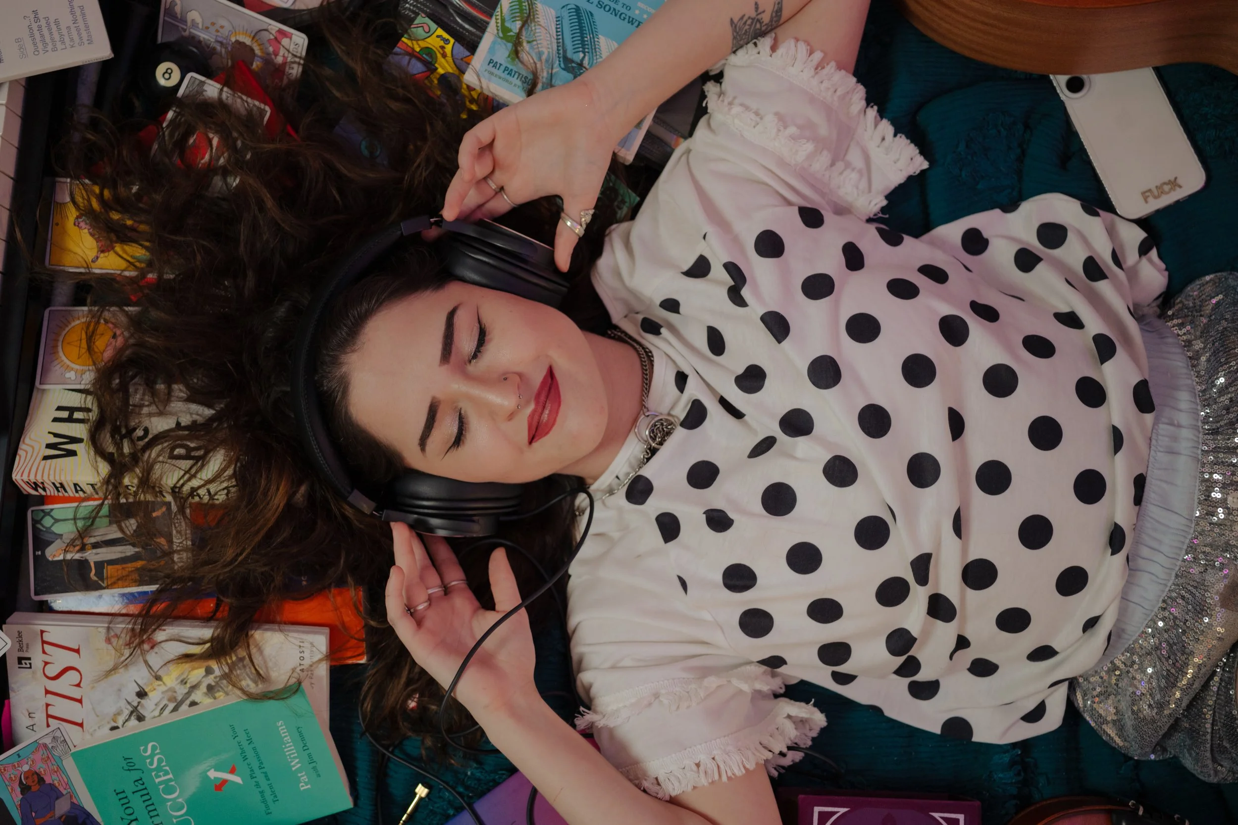 A young woman with dark curly hair, wearing headphones, a white polka dot shirt, and sparkly pants, lying on a surface surrounded by books and magazines.