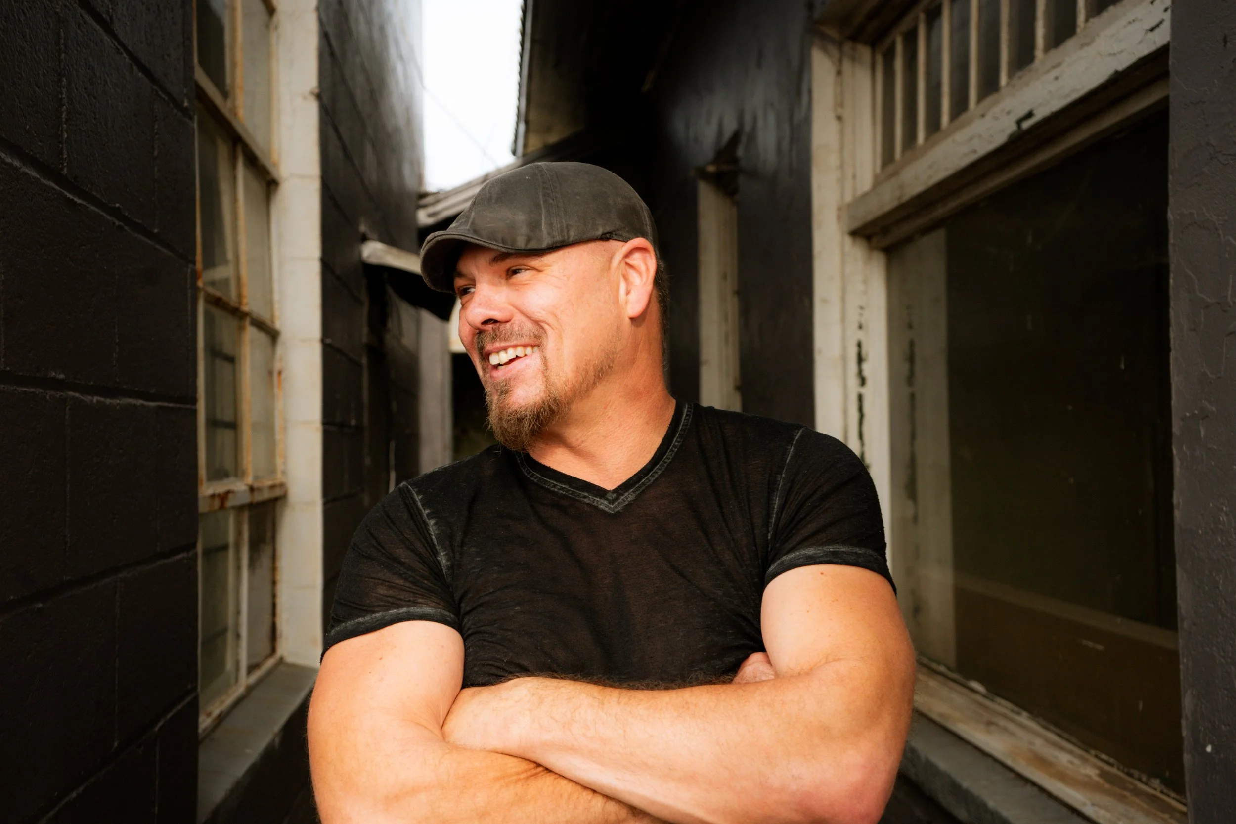 A smiling man with a beard and mustache wearing a black cap and black t-shirt, standing with arms crossed in an urban alleyway with black walls and windows.