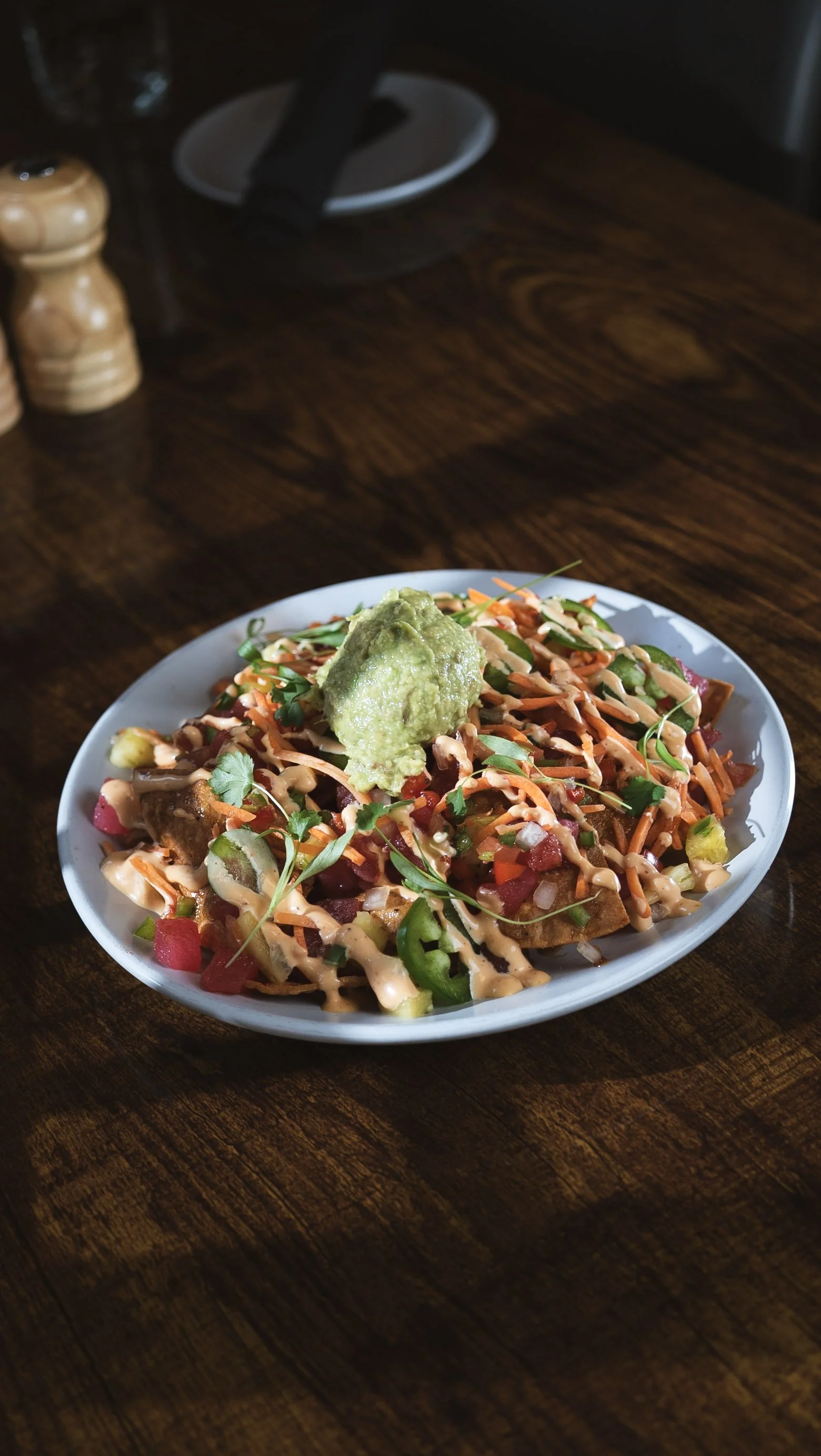 A plate of nachos topped with melted cheese, guacamole, diced tomatoes, jalapeños, cilantro, and drizzled with a creamy sauce, on a wooden table.