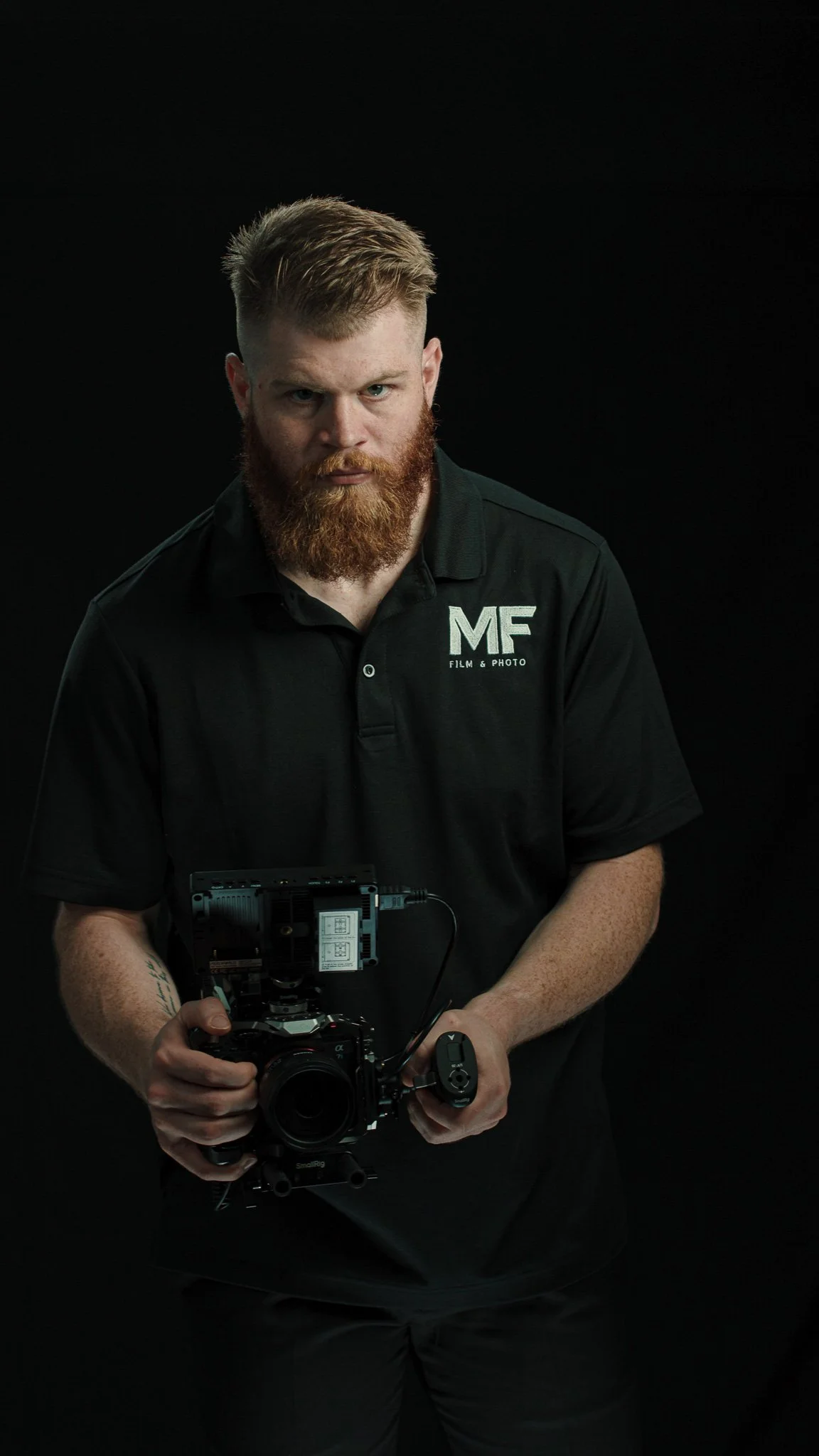 A man with red hair and a beard holding a professional camera against a black background. He is wearing a black polo shirt with 'MF Film & Photo' logo.