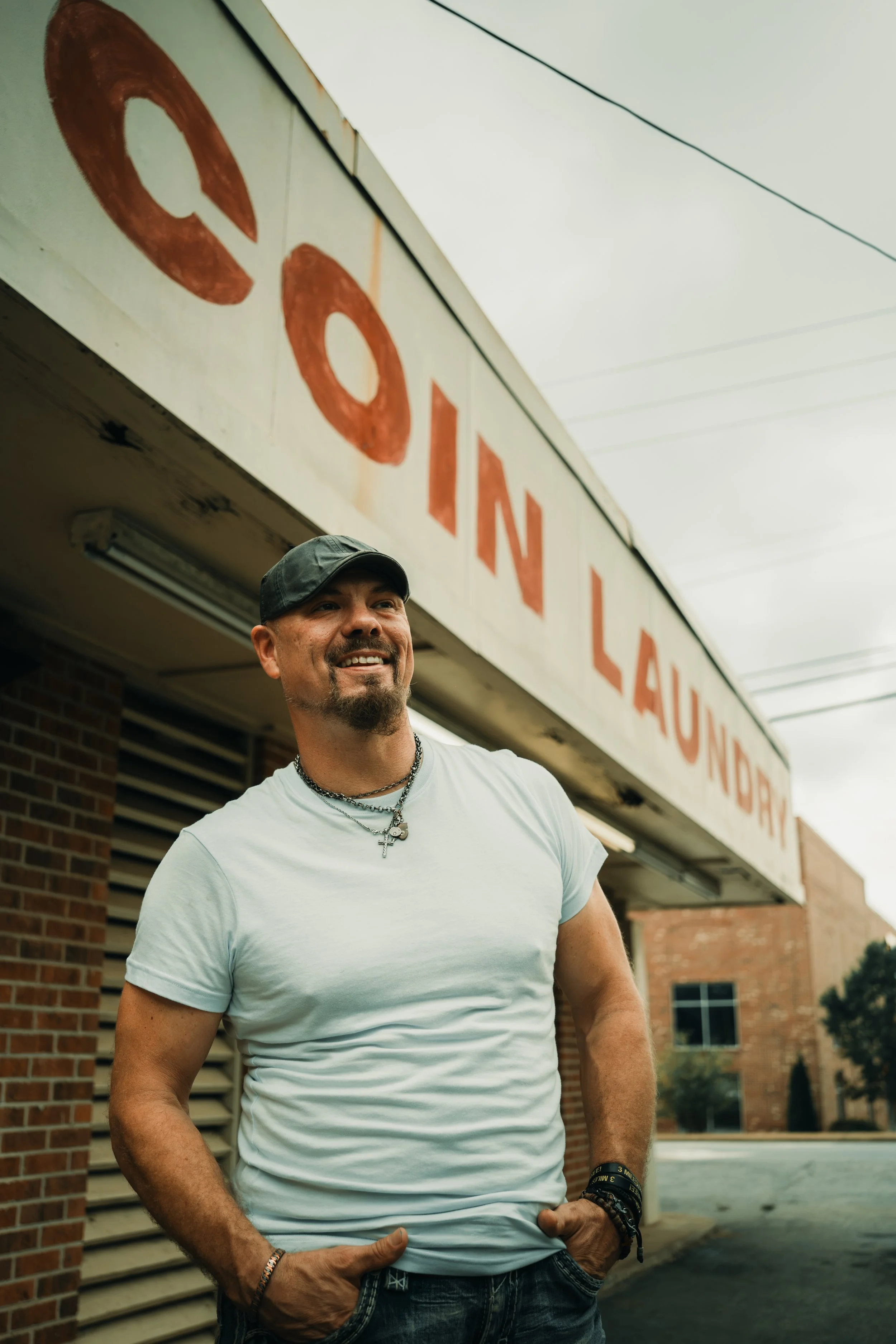 A man smiling, wearing a white t-shirt, jewelry, a black cap, and wristbands, standing outside near a brick building with a sign reading 'COIN LAUNDRY' in large letters.