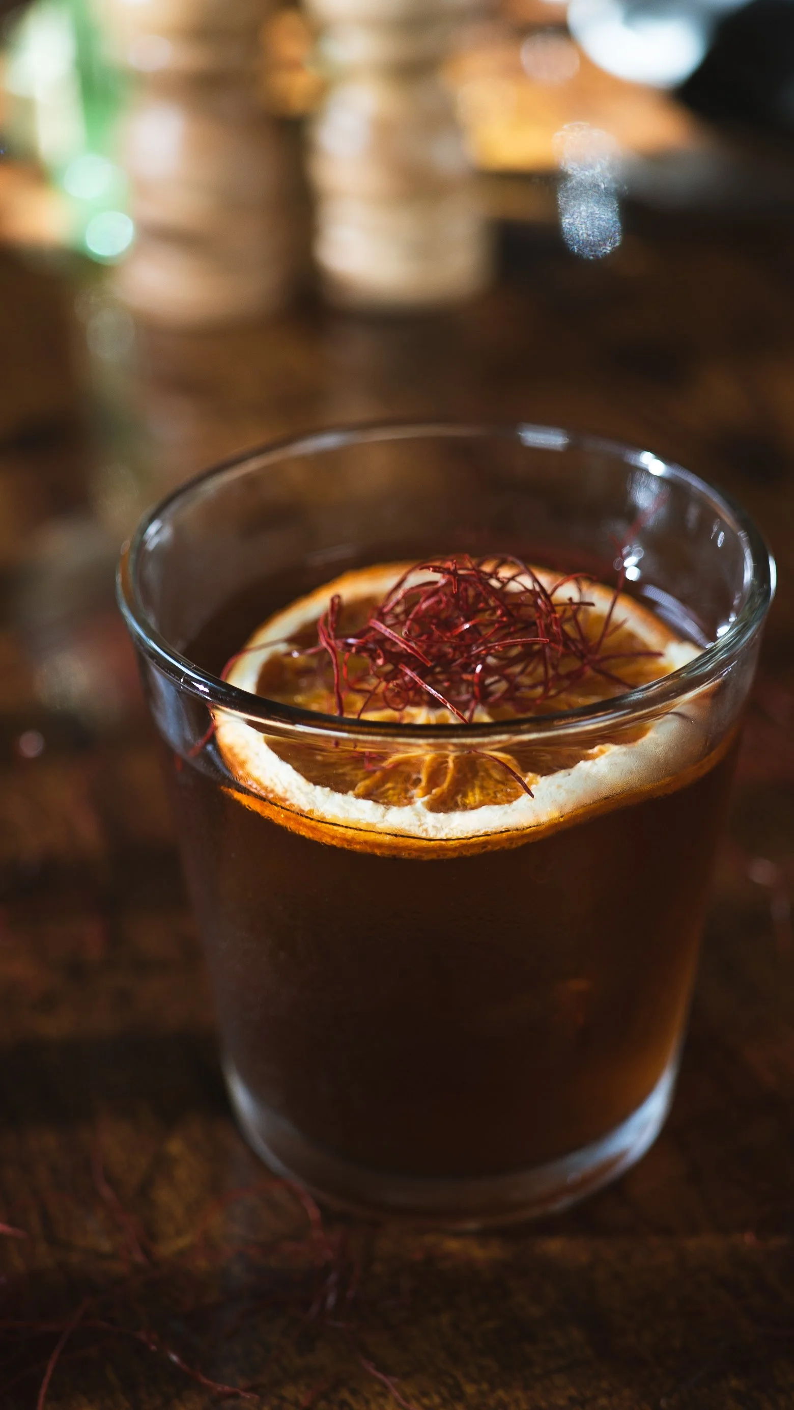 A glass of dark beverage garnished with a slice of orange and red thread-like herbs placed on top, sitting on a wooden surface.