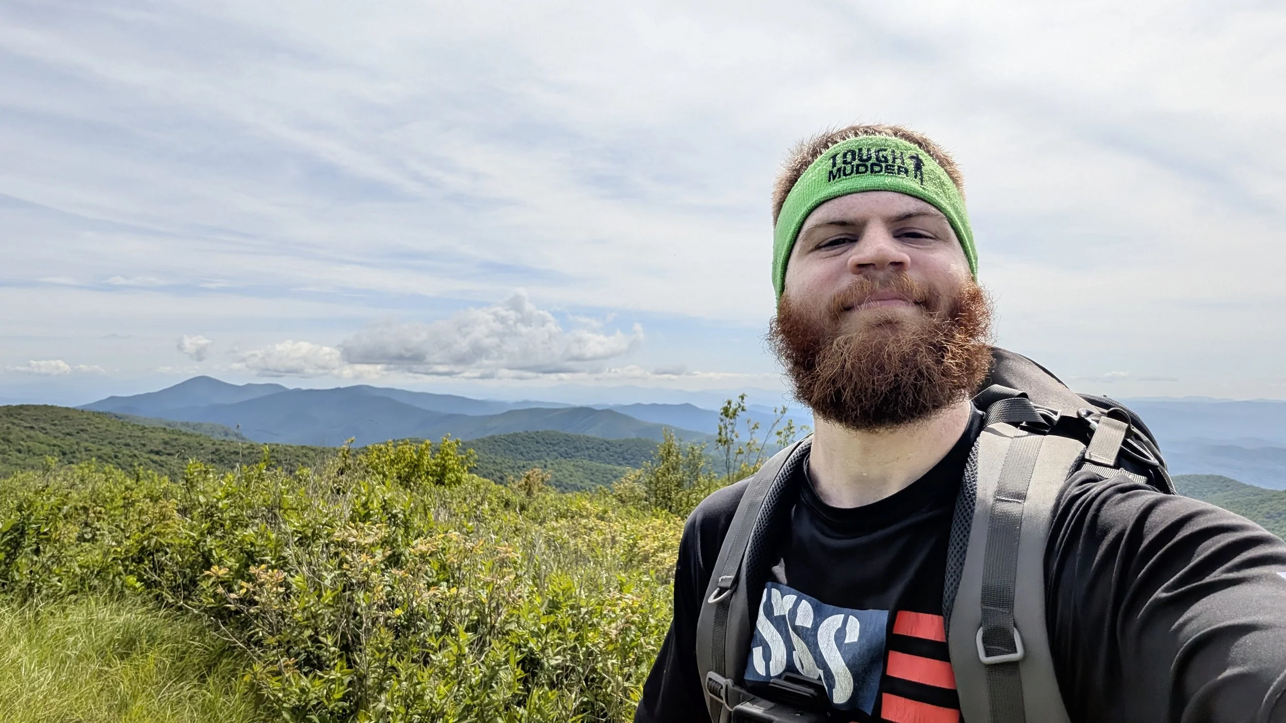 A man with a beard wearing a green headband and black shirt, taking a selfie on a mountain trail with green mountains and a cloudy sky in the background.