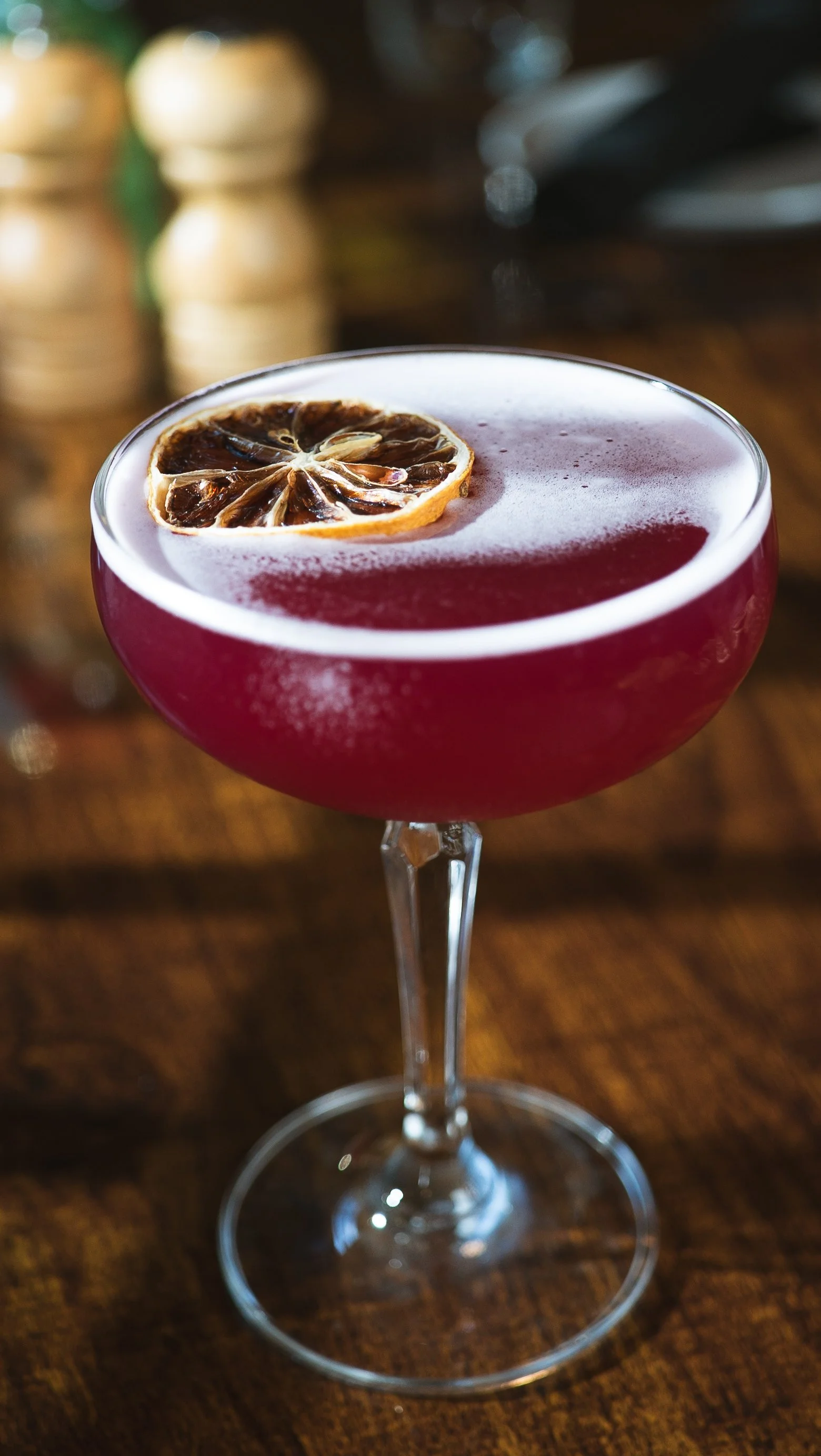 A cocktail in a stemmed glass with a dried blood orange slice garnish, placed on a wooden surface with a blurred background.