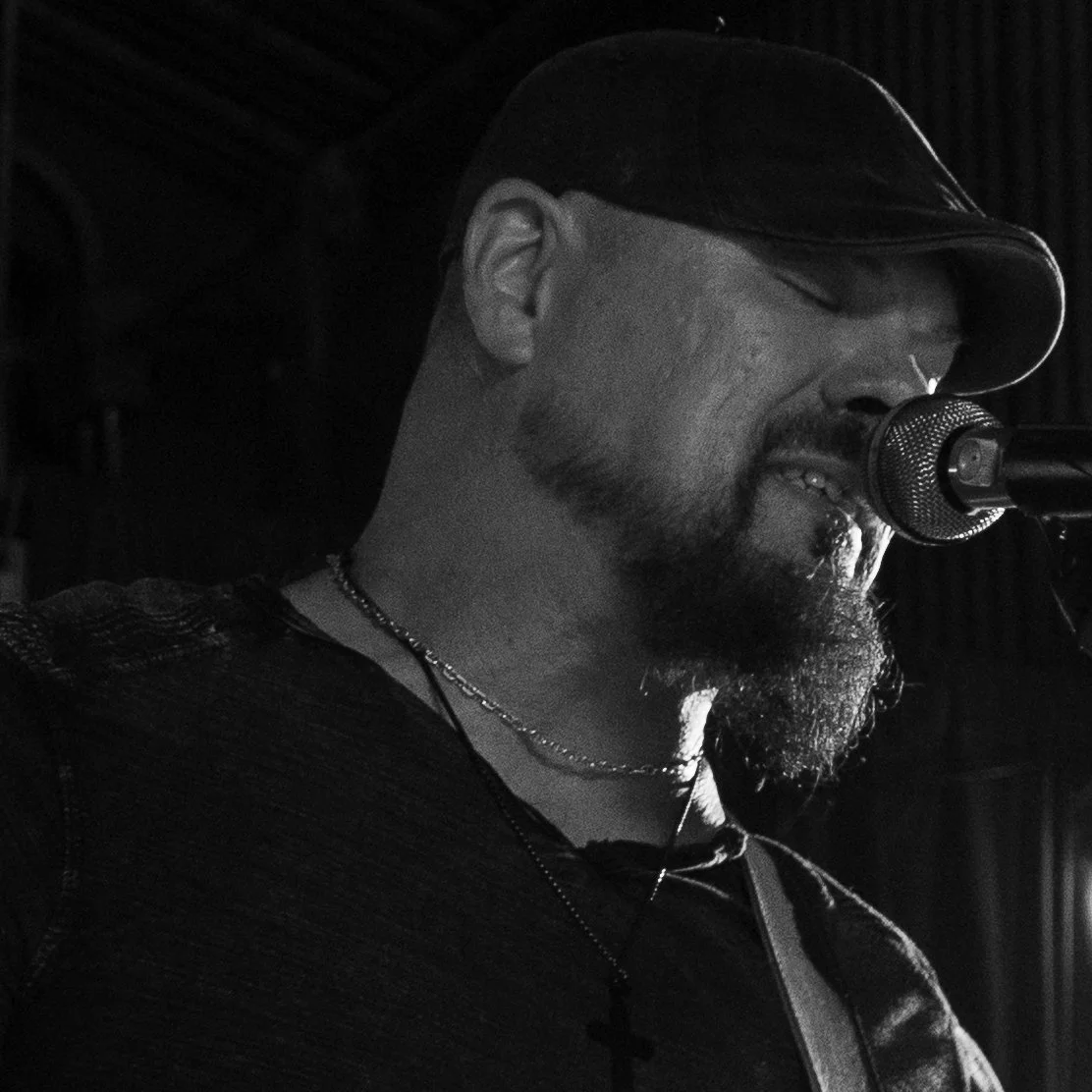 A man singing into a microphone, wearing a cap, glasses, and a necklace with a cross pendant, in a dark environment