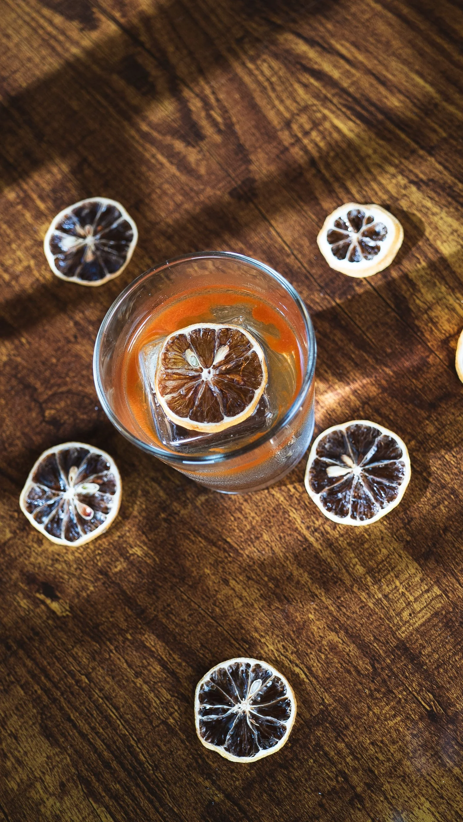 A glass of beverage garnished with a lemon slice, on a wooden table surrounded by dried lemon slices.