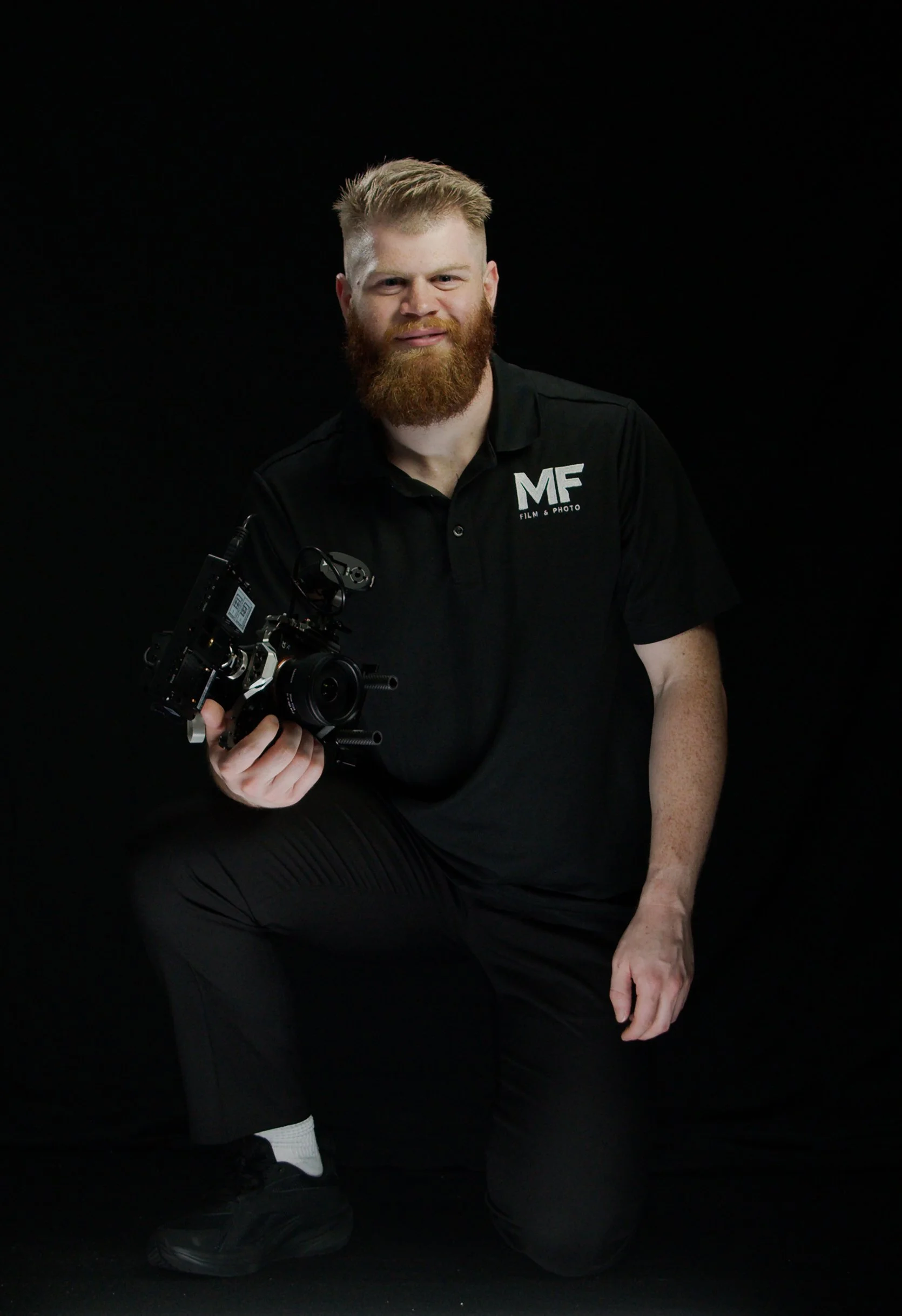 A man with a blonde haircut and a red beard is holding a professional camera. He is wearing a black polo shirt with the text 'MF Film & Photo' on it, black pants, and black shoes. The background is black.