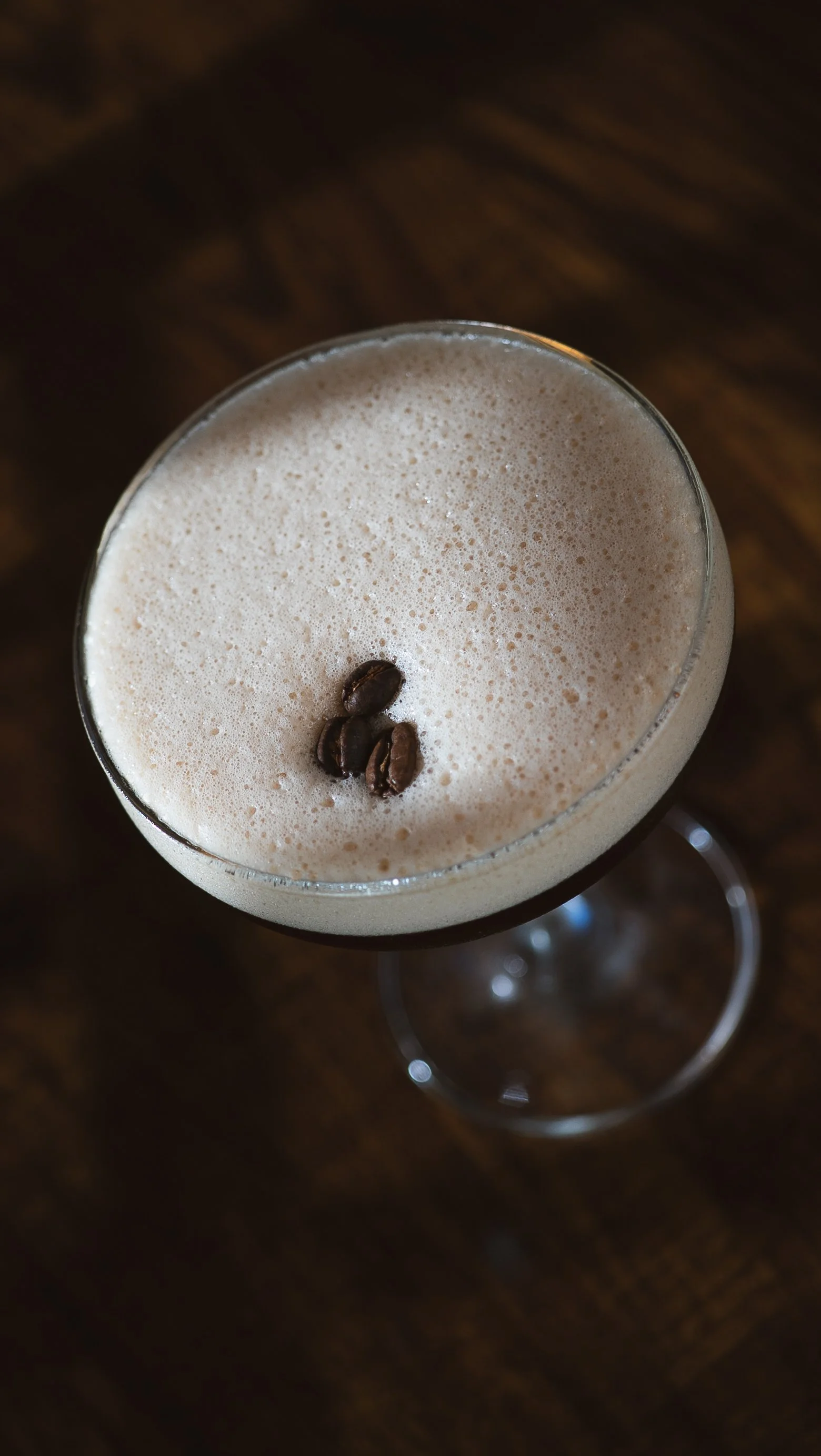 A glass of coffee with foam and three coffee beans on top, placed on a dark wooden surface.