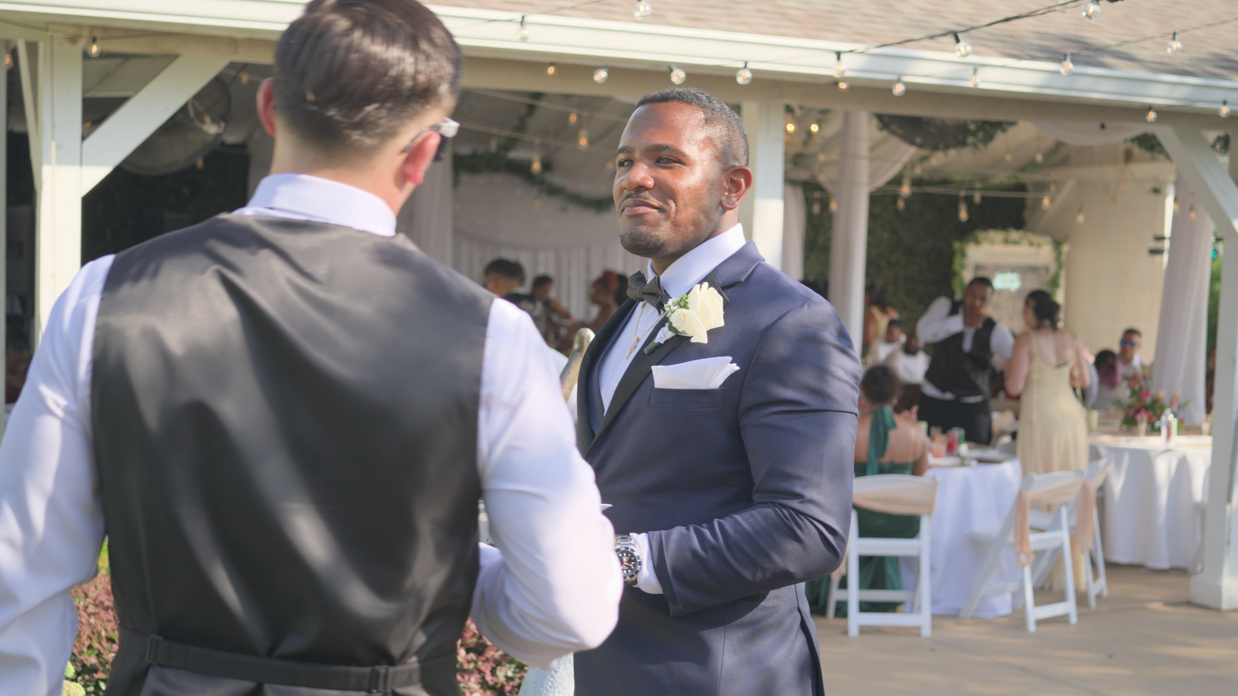 Two men in formal attire laughing at a wedding reception outdoors, with guests seated at decorated tables in the background.