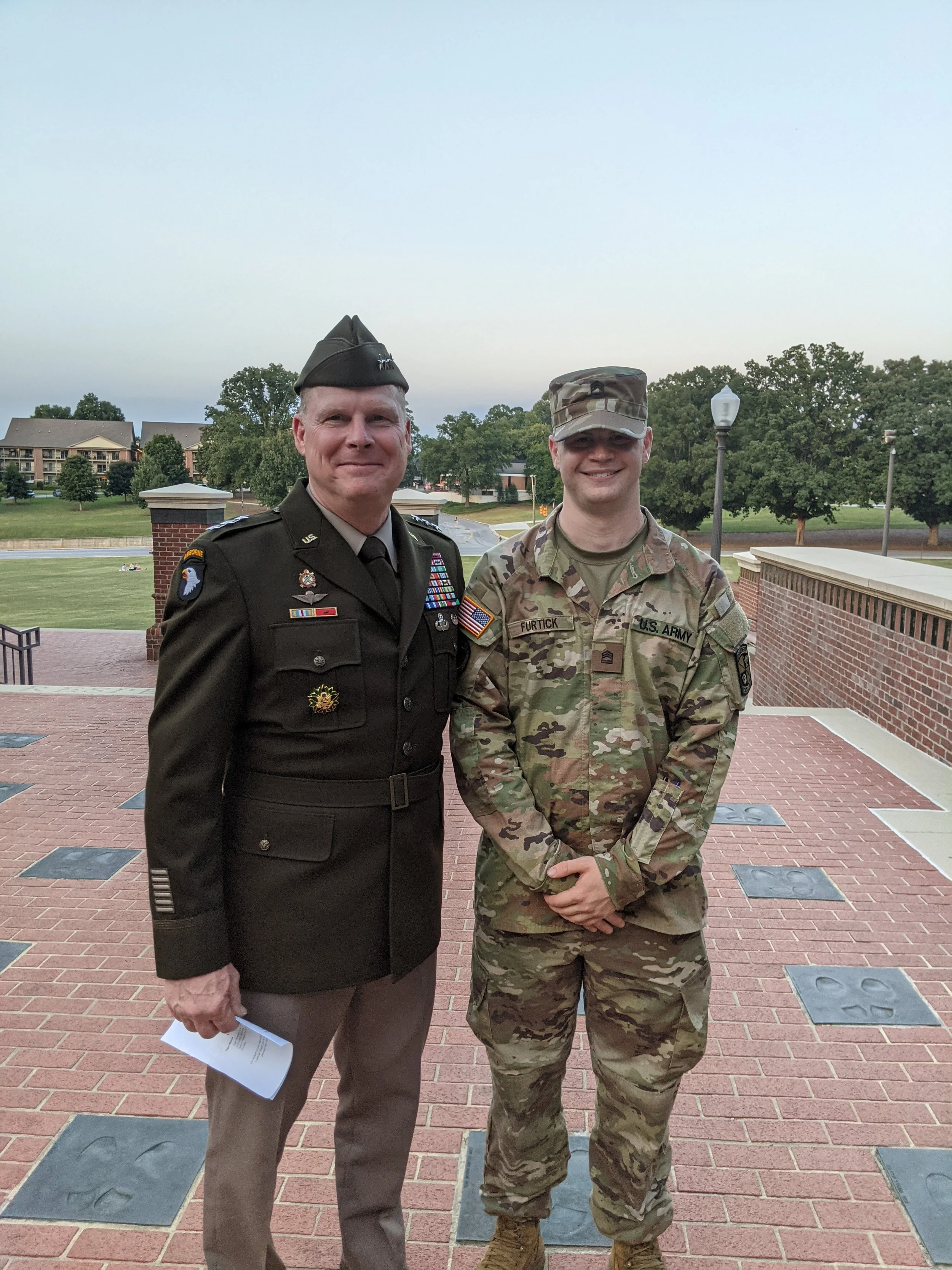 Two men in military uniform standing outside on a brick pathway with trees and lamp posts in the background.