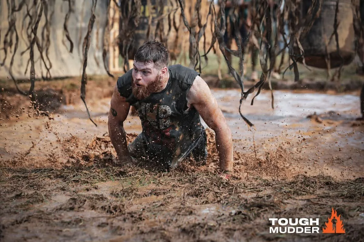 A man with a beard and athletic build crawling through muddy water during an obstacle course, with the "Tough Mudder" logo in the bottom right corner.