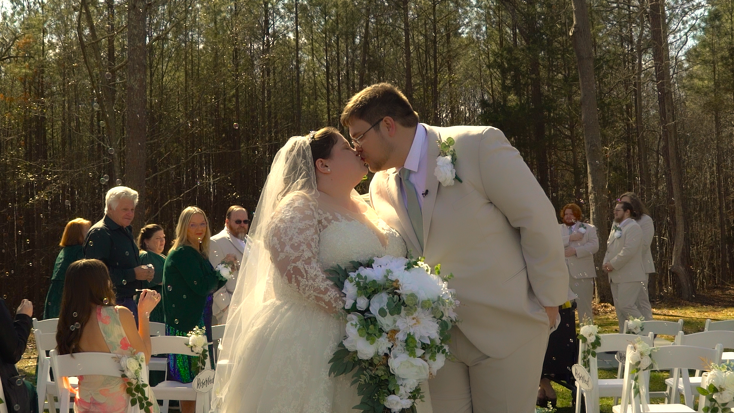 A bride and groom share a kiss at an outdoor wedding ceremony in a wooded area, with guests in the background.
